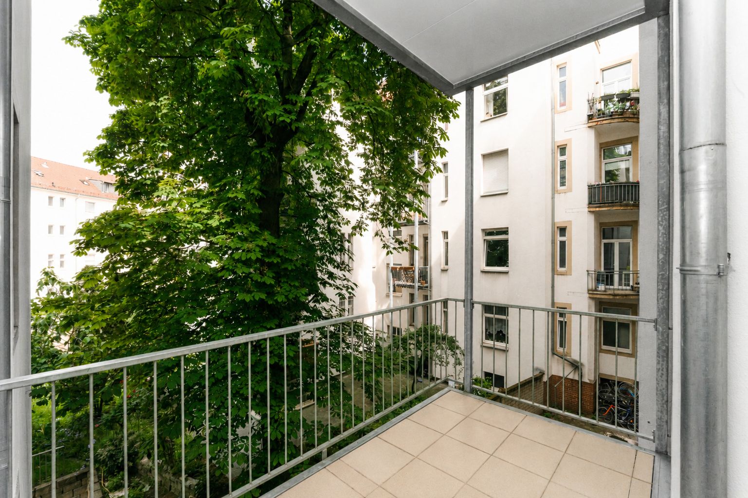 Überdachter Balkon mit Metallgeländer, Blick auf Baum und benachbarte Wohnhäuser.