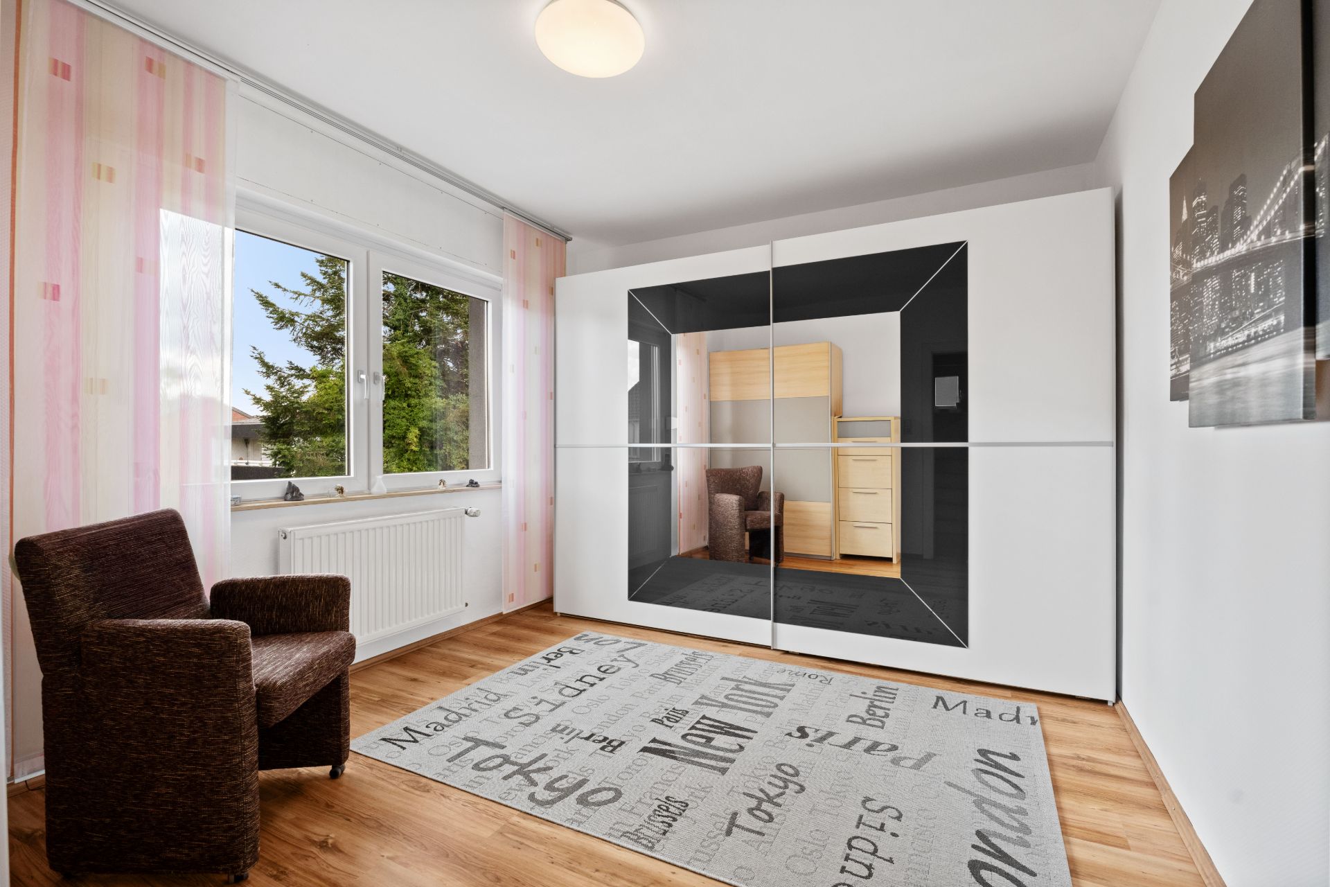 Helles Zimmer mit Sessel, großem Kleiderschrank, Teppich und Fenster mit Blick nach draußen.