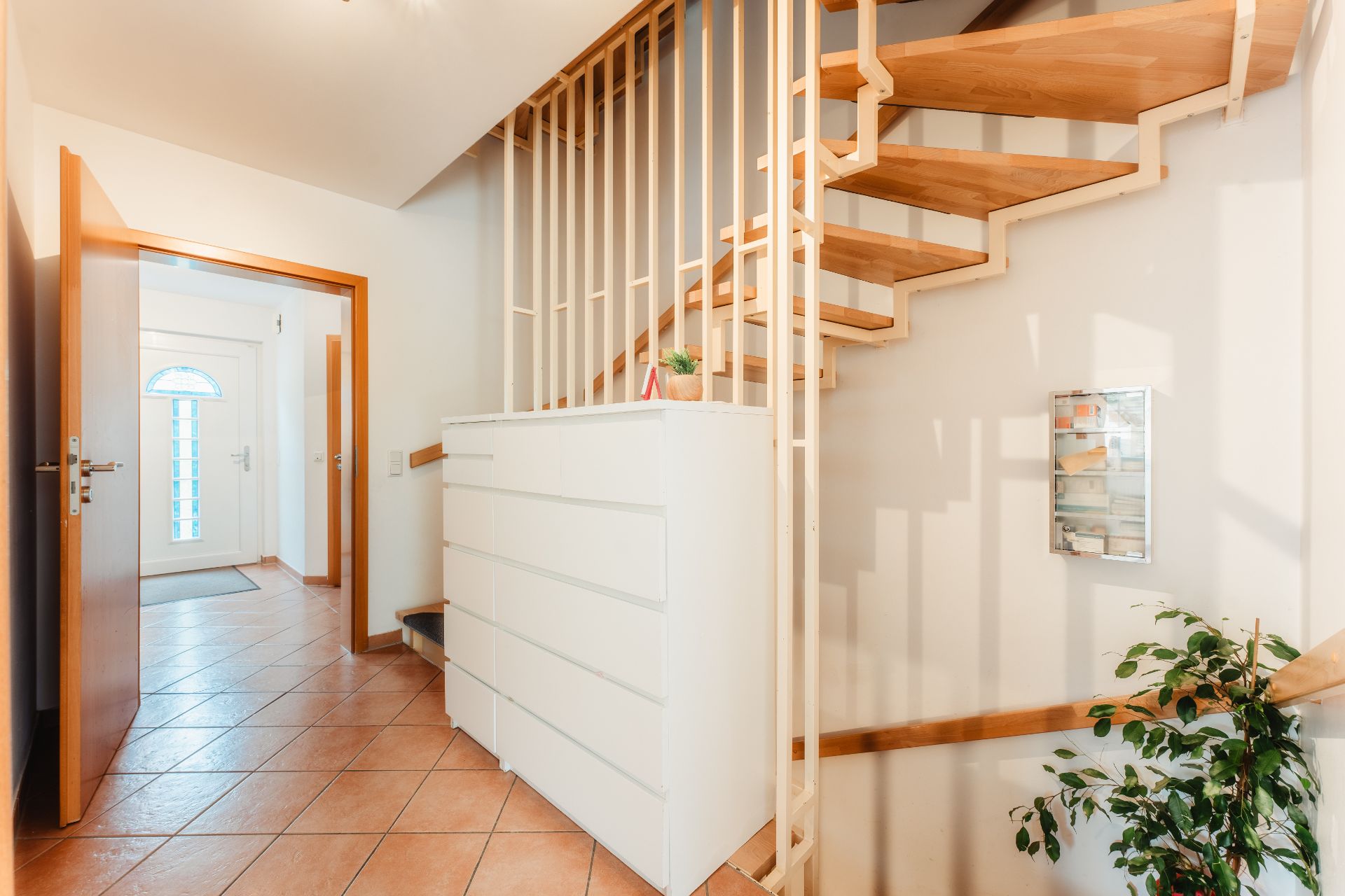 Heller Flur mit Treppe aus Holz, Kommode, Pflanze und Eingangstür im Hintergrund.