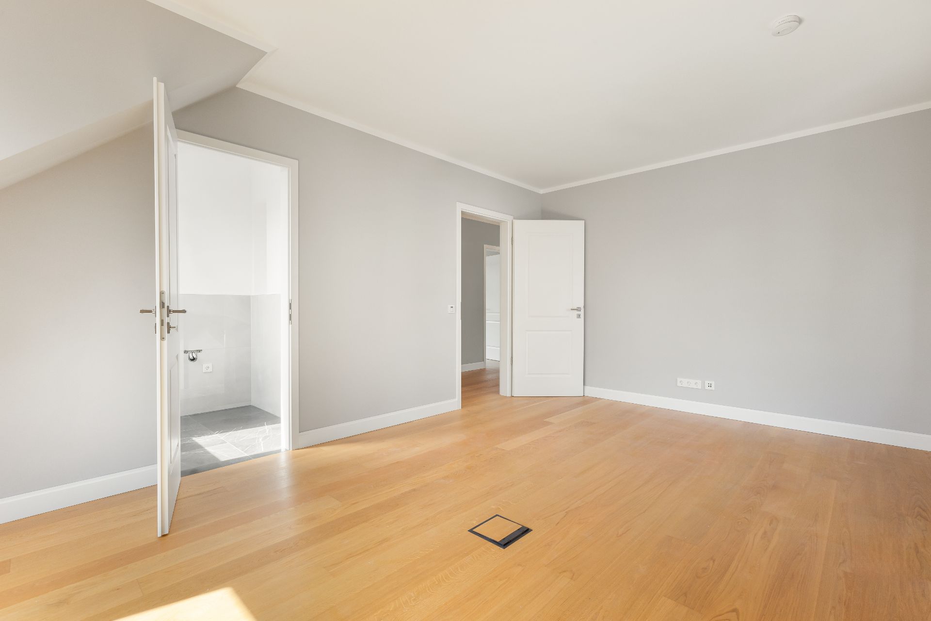 Leerer Raum mit Holzboden, zwei offenen Türen und grauen Wänden.