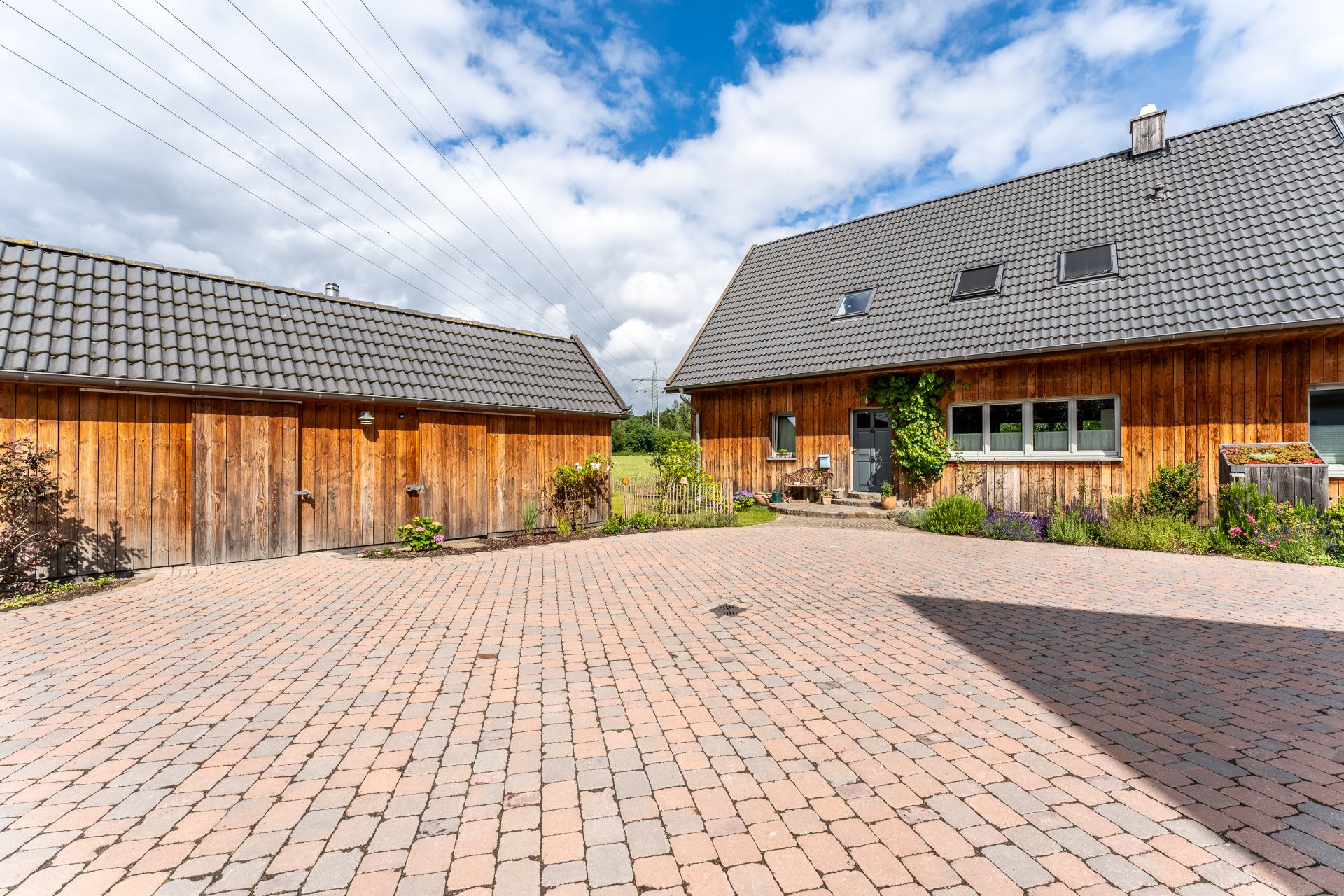 Gepflasterter Hof mit zwei Gebäuden aus Holz und gepflegten Pflanzen am Rand.