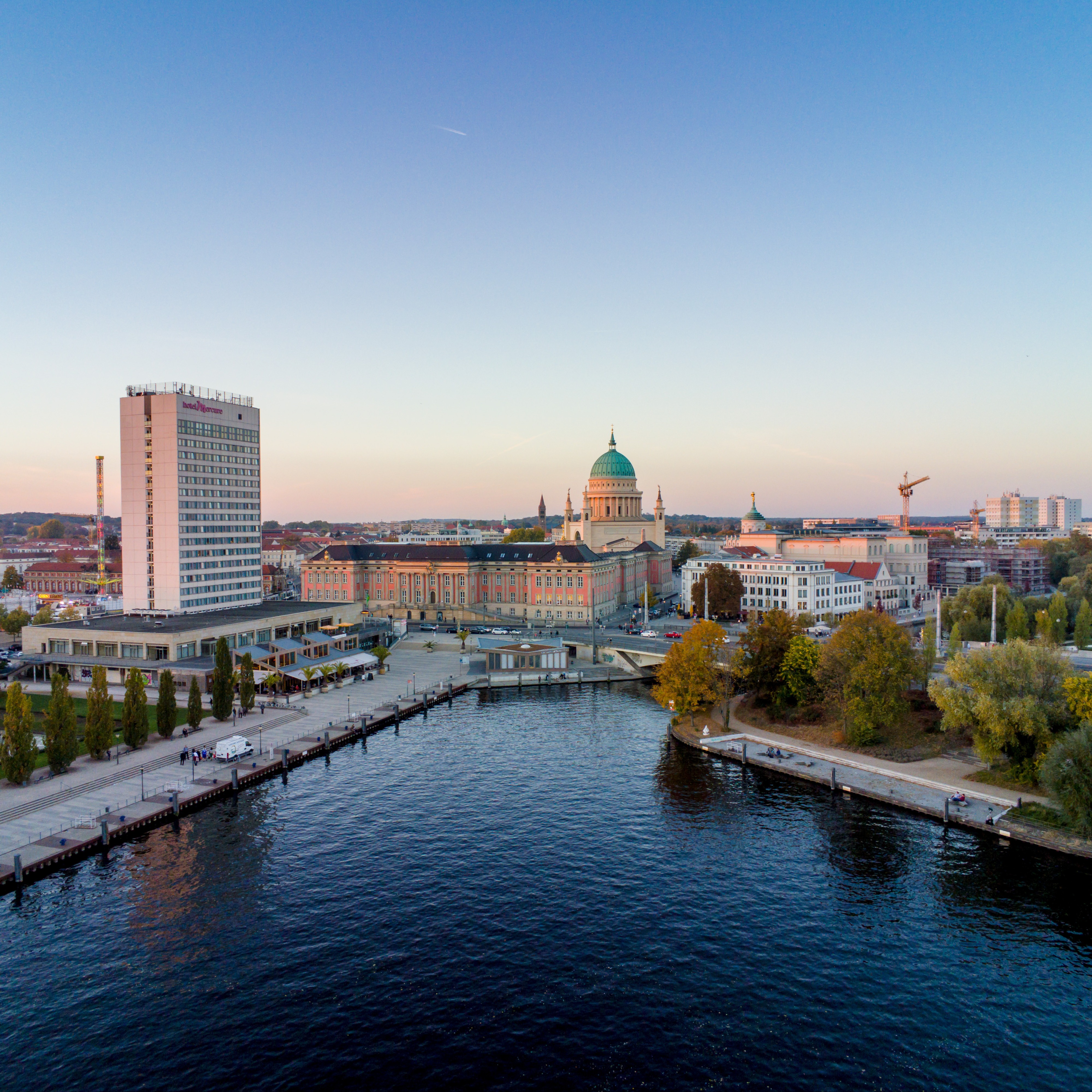 Stadtansicht mit Fluss, modernen und historischen Gebäuden sowie einer Uferpromenade.
