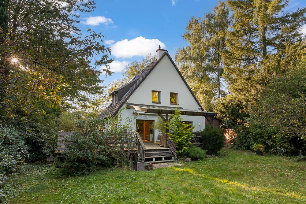 Weißes Einfamilienhaus mit spitzem Dach und Holzterrasse im grünen Garten.