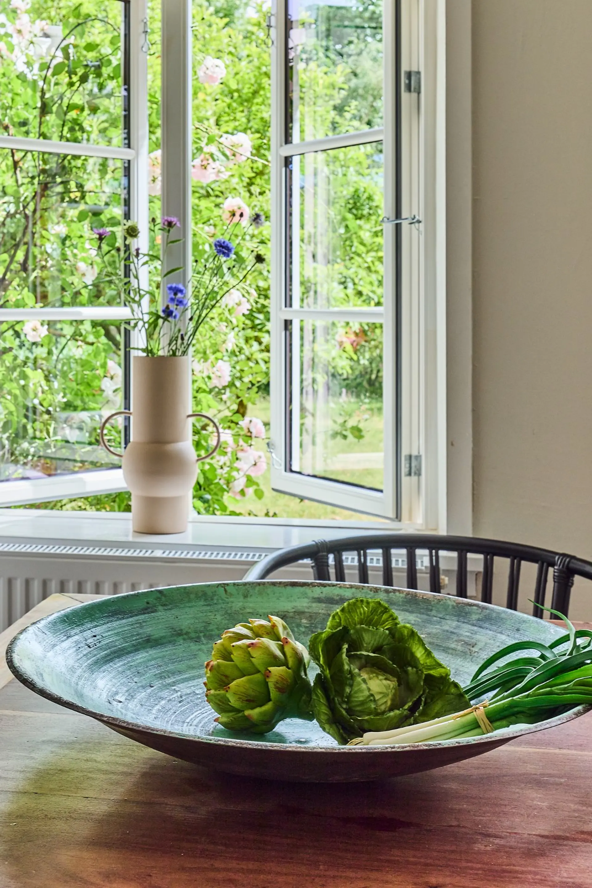 Offenes Fenster mit Blick ins Grüne, Vase mit Blumen auf der Fensterbank, Schüssel mit Gemüse auf dem Tisch.