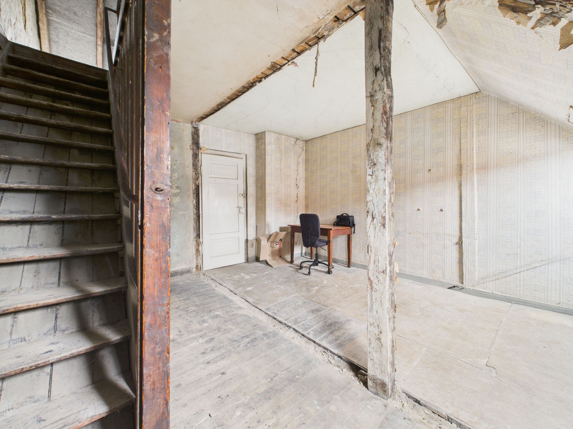 Ein veralteter Raum mit einer Holztreppe, einem Schreibtisch und einer Stuhlsäule, teils renovierungsbedürftig.