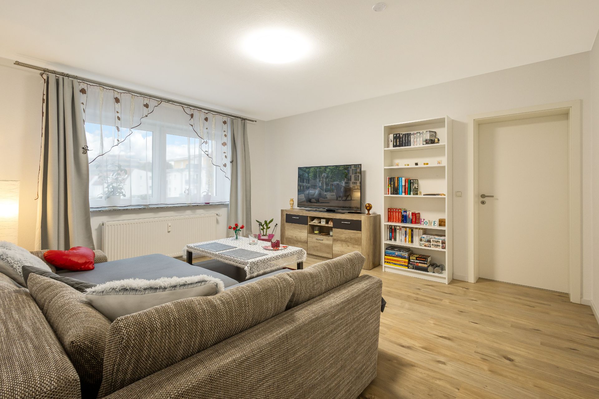 Helles Wohnzimmer mit grauem Sofa, Couchtisch, Fernseher und Bücherregal auf Holzfußboden.