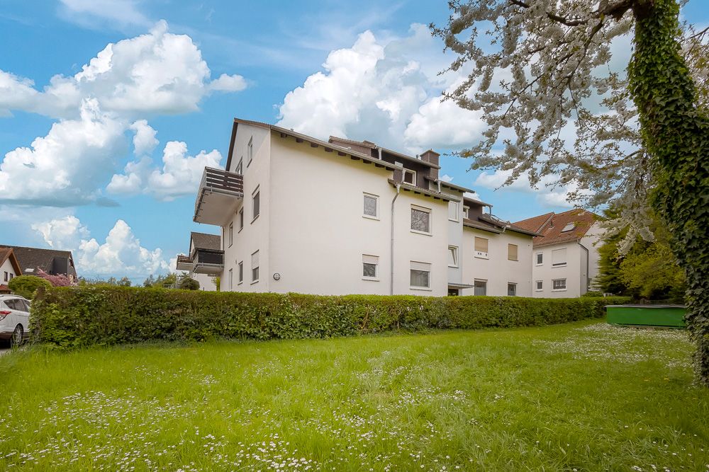 Mehrfamilienhaus mit Balkonen, umgeben von einer grünen Wiese und Bäumen an einem sonnigen Tag.