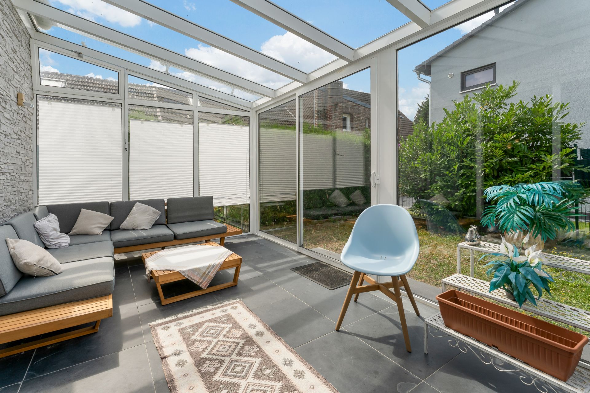 Heller Wintergarten mit Ecksofa, Stuhl, Pflanzen und Glastüren zum Garten.