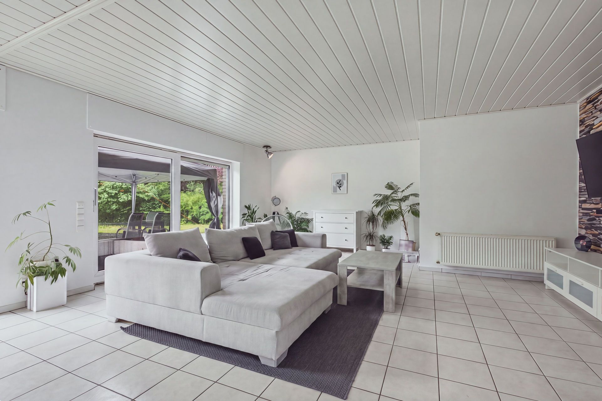 Helles Wohnzimmer mit Ecksofa, Couchtisch, Pflanzen und Zugang zur Terrasse.
