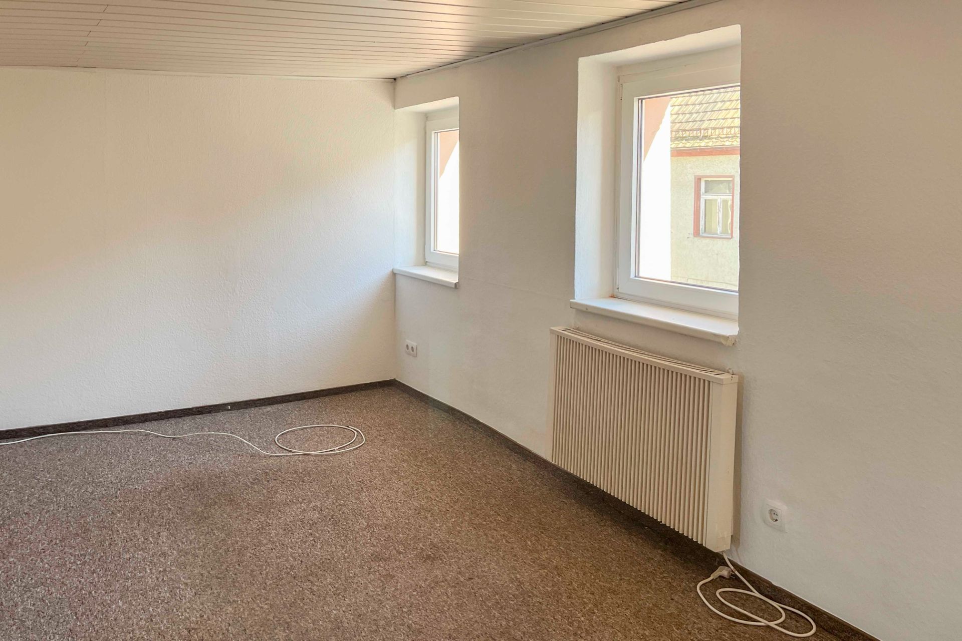 Leeres Zimmer mit zwei Fenstern, einem Heizkörper und Teppichboden.