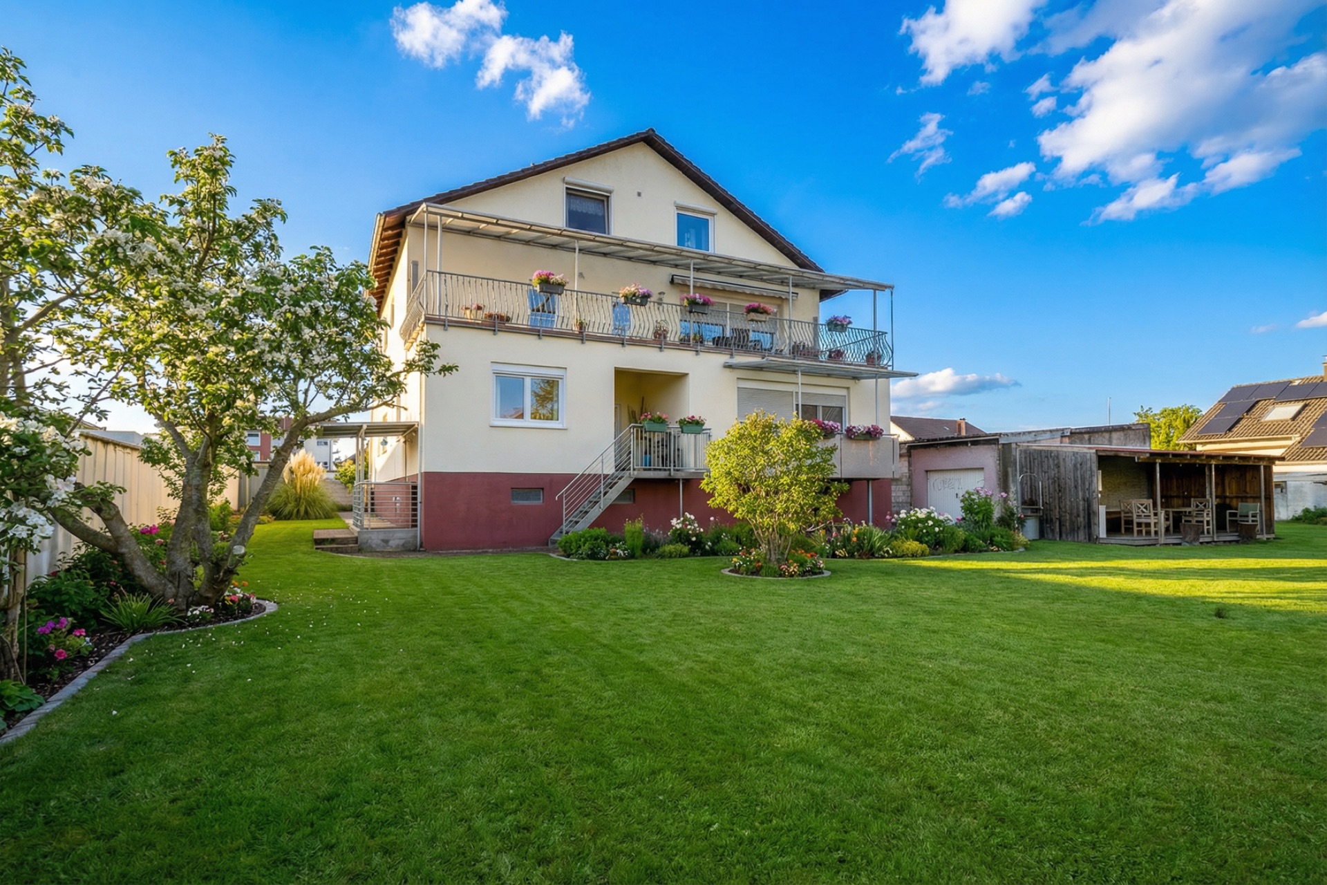 Zweistöckiges Wohnhaus mit Balkon, Garten, Bäumen und überdachtem Sitzbereich im Hof.