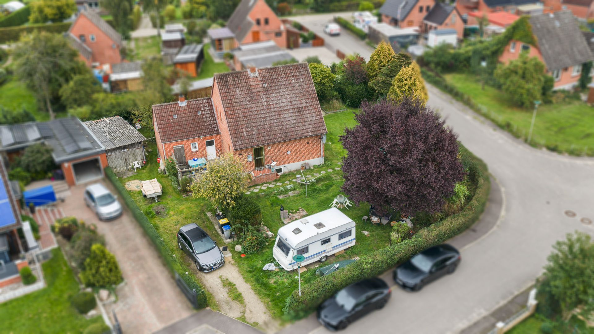 Luftaufnahme eines Einfamilienhauses mit Garten, Wohnwagen und geparkten Autos an einer Kurve.