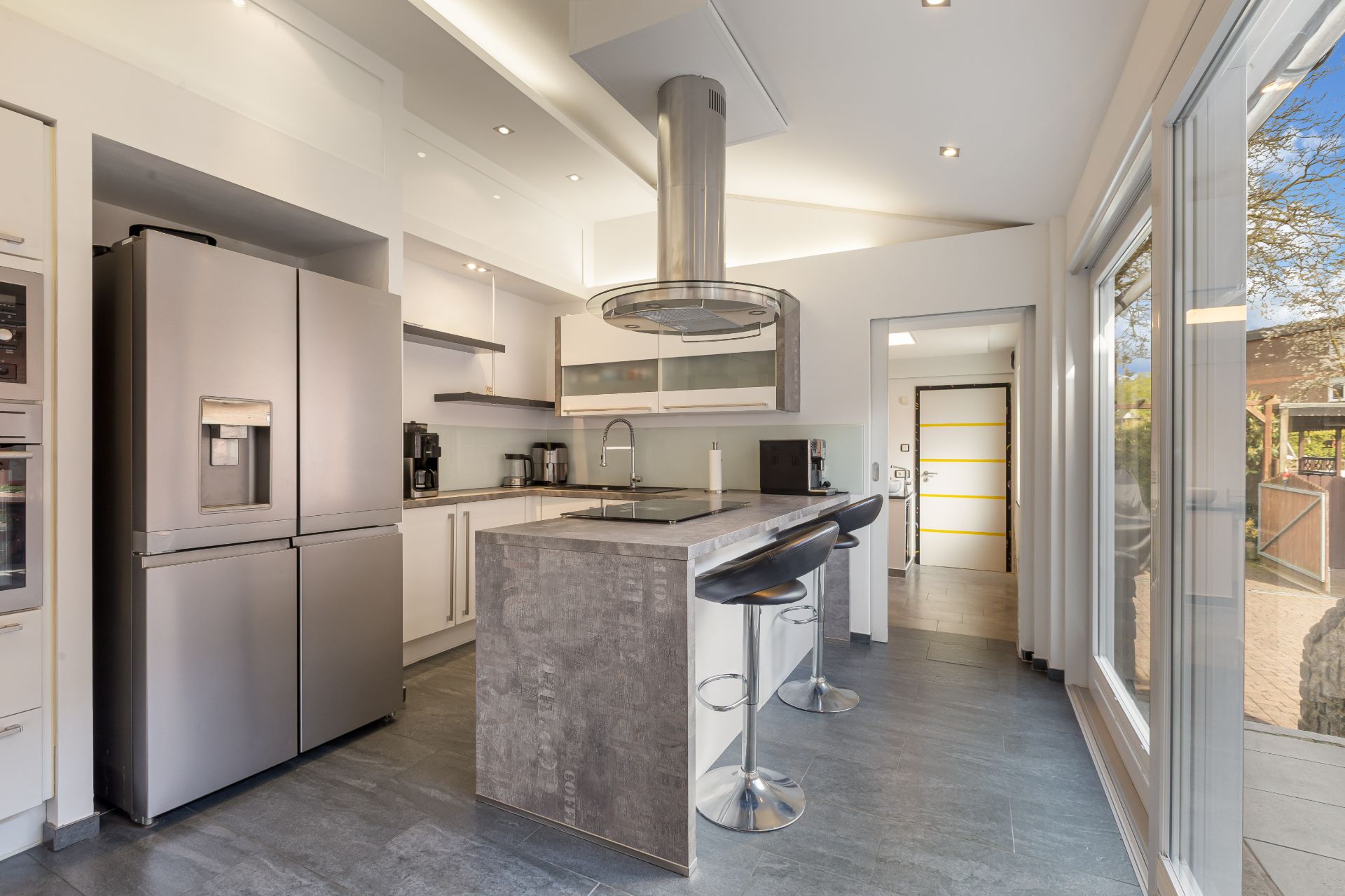 Moderne Küche mit Kochinsel, Barhockern, großem Kühlschrank und bodentiefen Fenstern.
