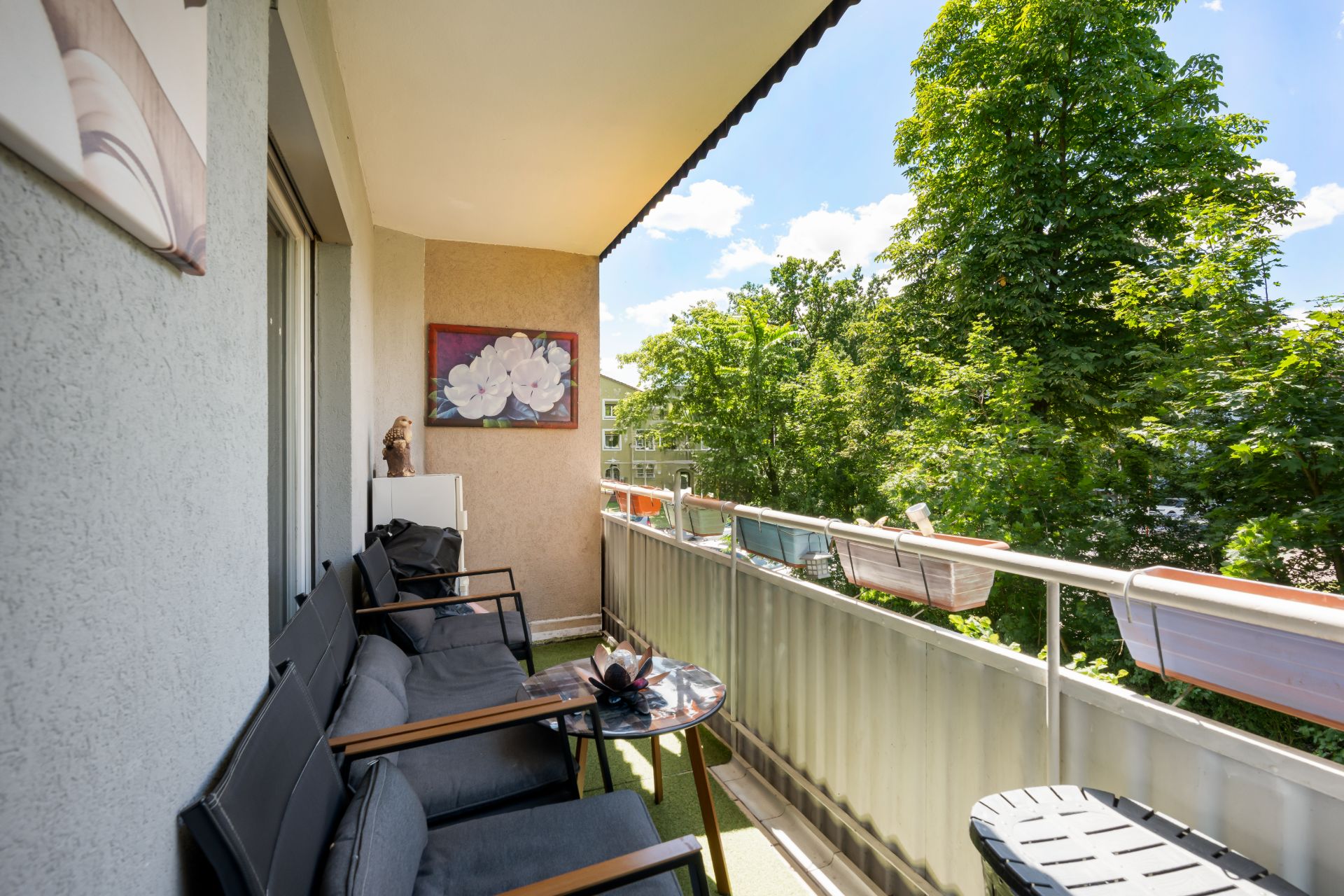 Balkon mit Sitzmöbeln, Tisch und Blick auf grüne Bäume an einem sonnigen Tag.