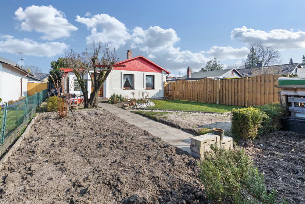 Kleines Haus mit rotem Dach und Garten, Weg führt durch das Grundstück, Bäume und Beete sichtbar.