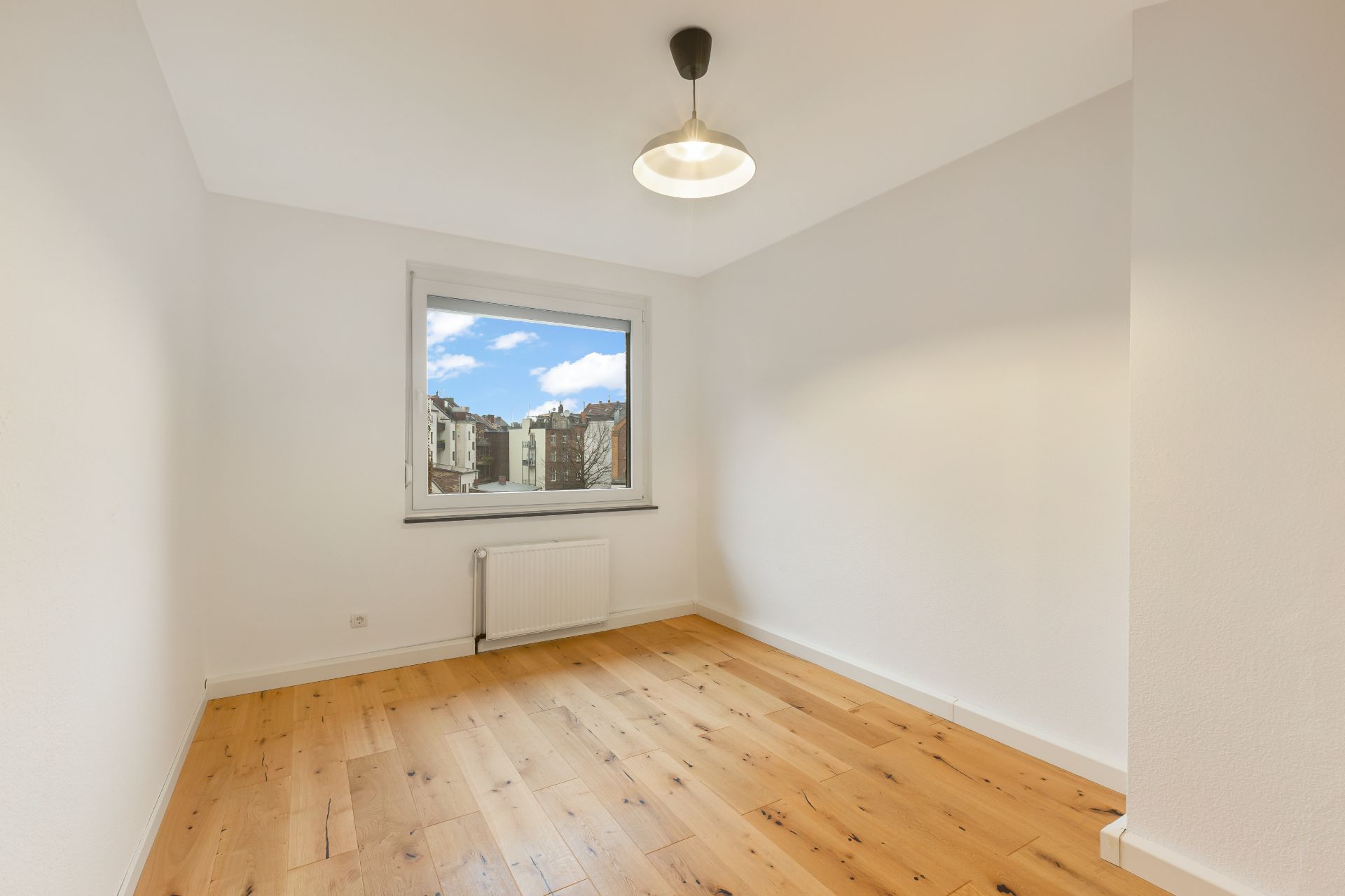 Helles, leeres Zimmer mit Holzboden, Fenster und Deckenlampe.