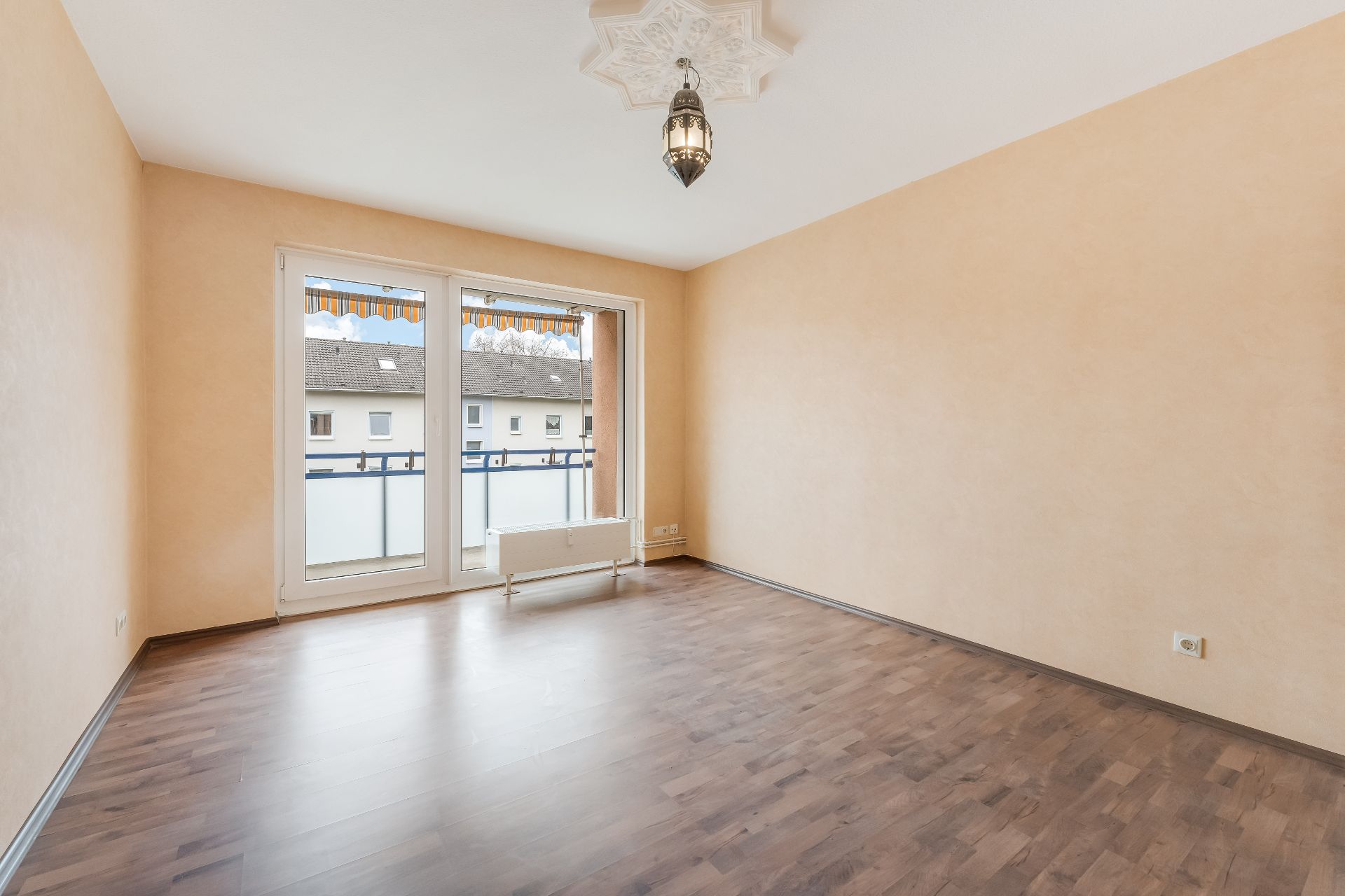 Leeres Zimmer mit Holzfußboden, beigen Wänden, großem Fenster und moderner Deckenleuchte.