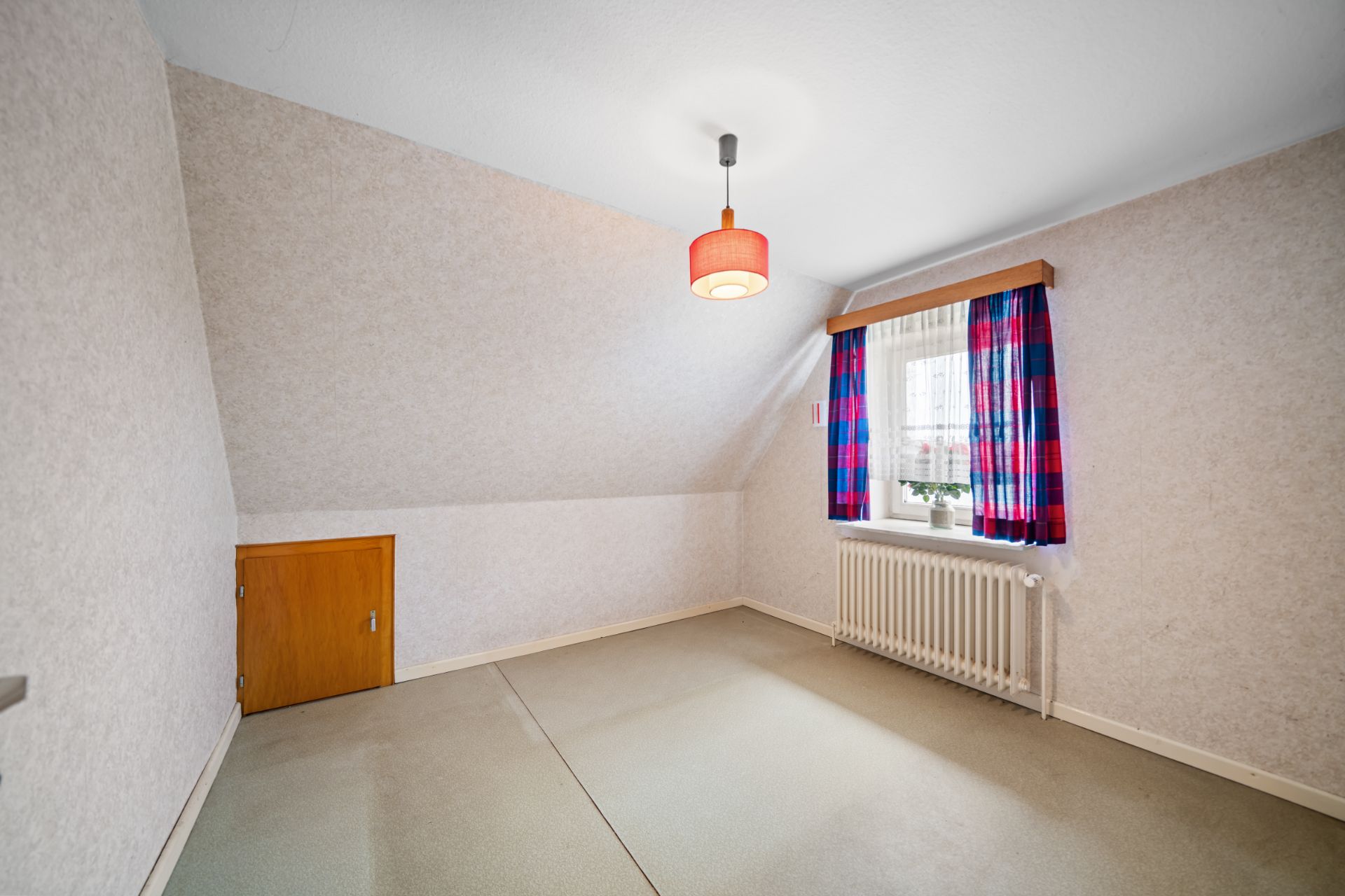 Leeres Zimmer mit schräger Decke, kleinem Fenster mit Vorhängen und Heizkörper darunter.