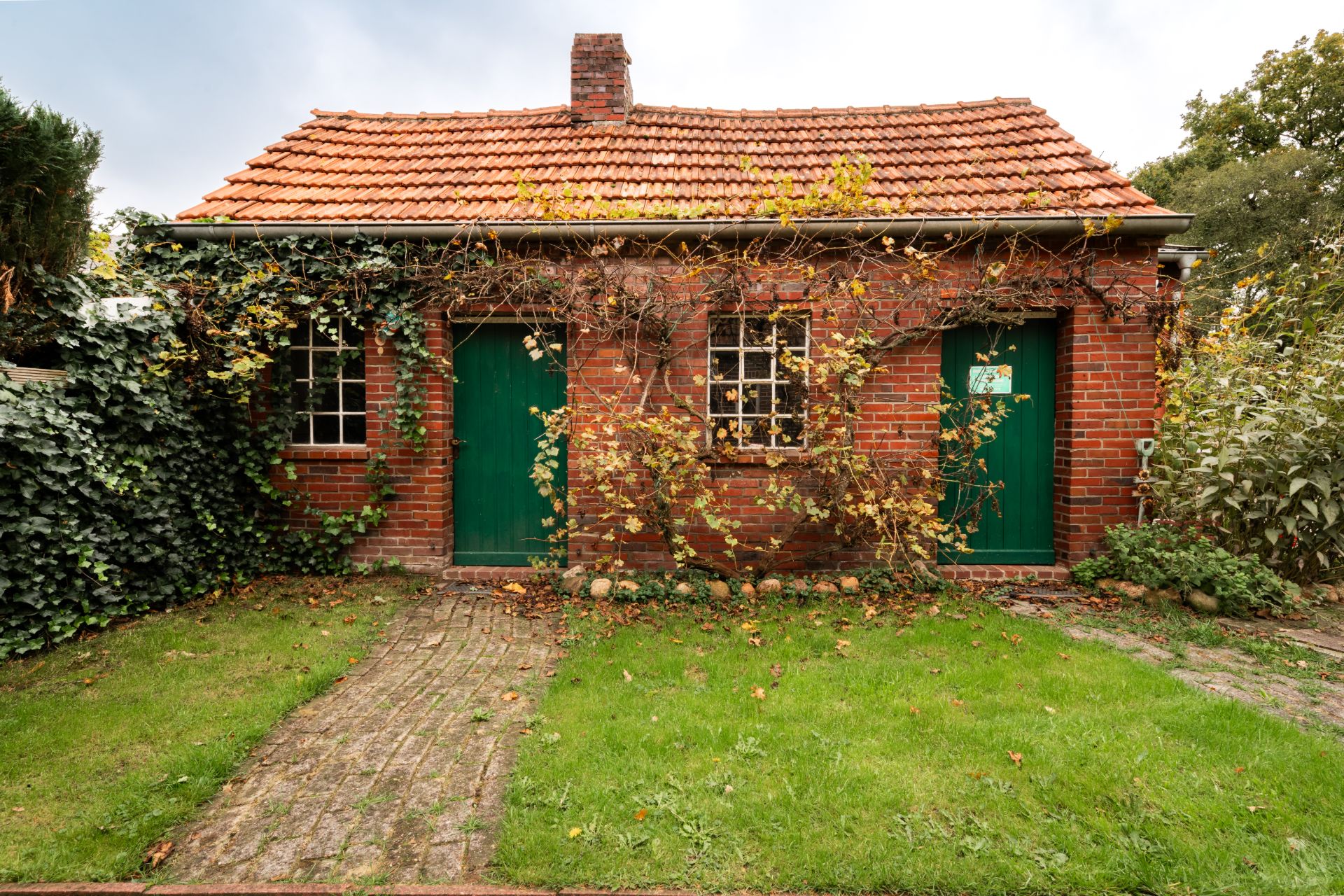 Backsteinhaus mit rotem Ziegeldach, zwei grünen Türen und überwachsener Fassade im Garten.