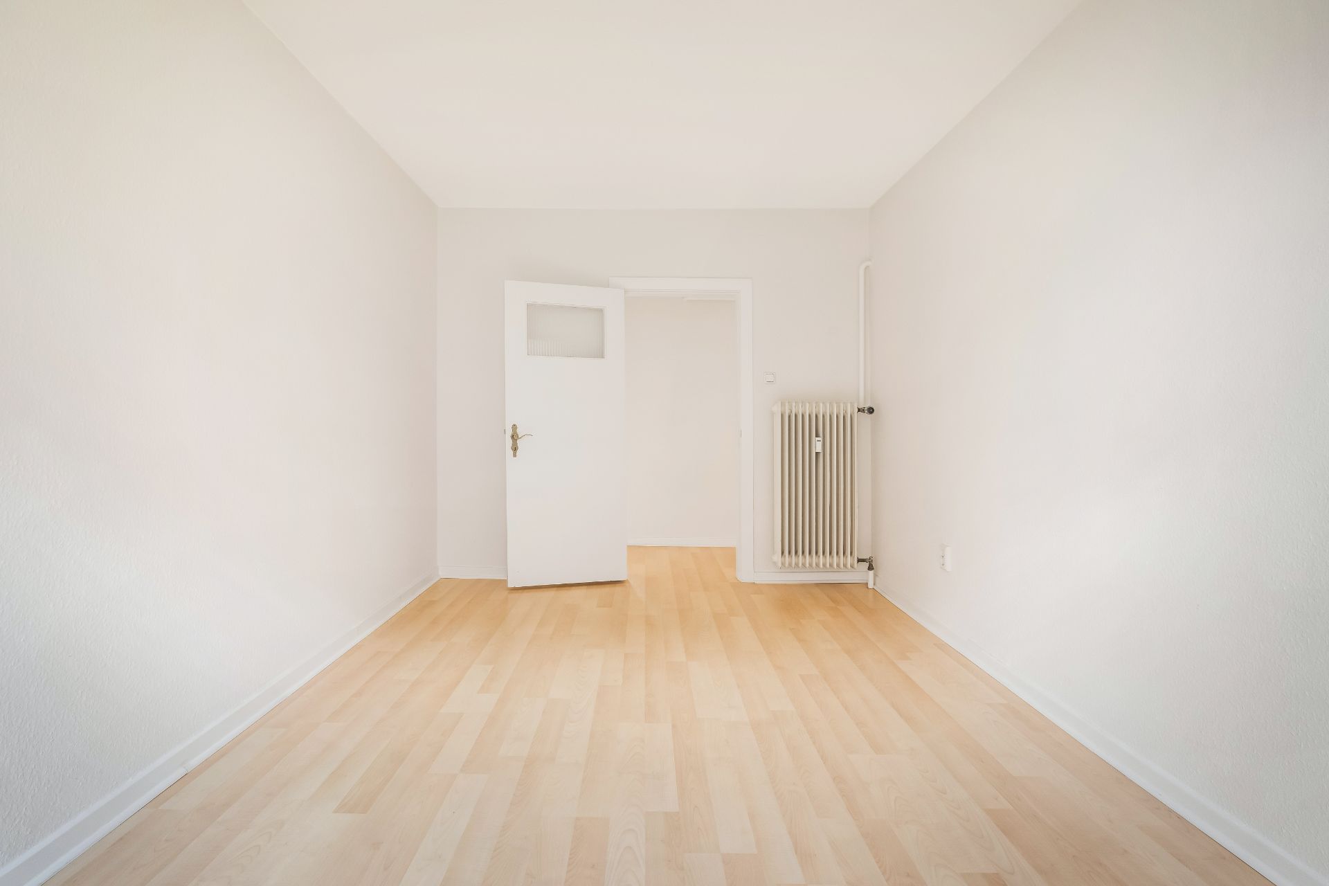 Leerer, heller Raum mit Holzfußboden, weißer Tür und Heizkörper an der Wand.