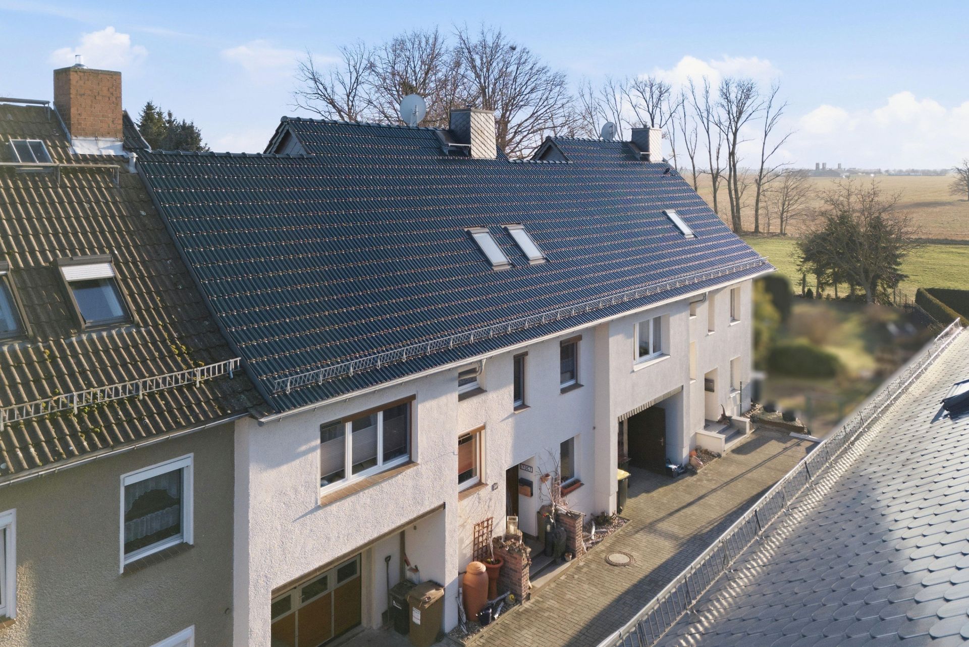 Mehrfamilienhaus mit mehreren Eingängen und dunklem Dach, dahinter Wiese und Bäume.
