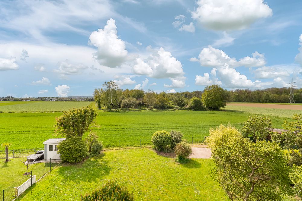 Blick auf einen gepflegten Garten mit Rasen, Bäumen und Gartenhaus, dahinter weite grüne Felder.