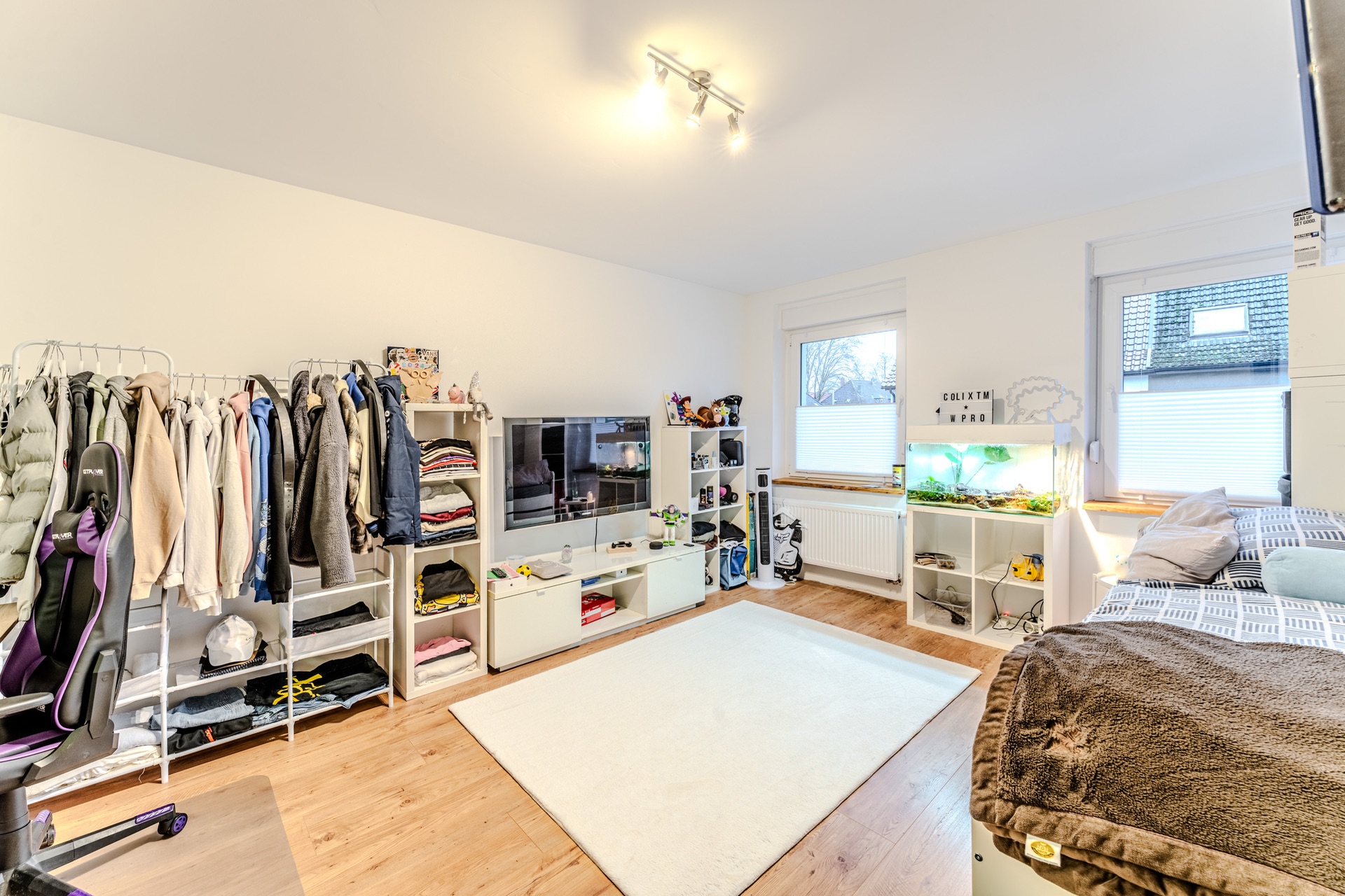 Helles Zimmer mit Bett, Fernseher, Kleiderständern, Aquarium und Teppich auf Holzboden.