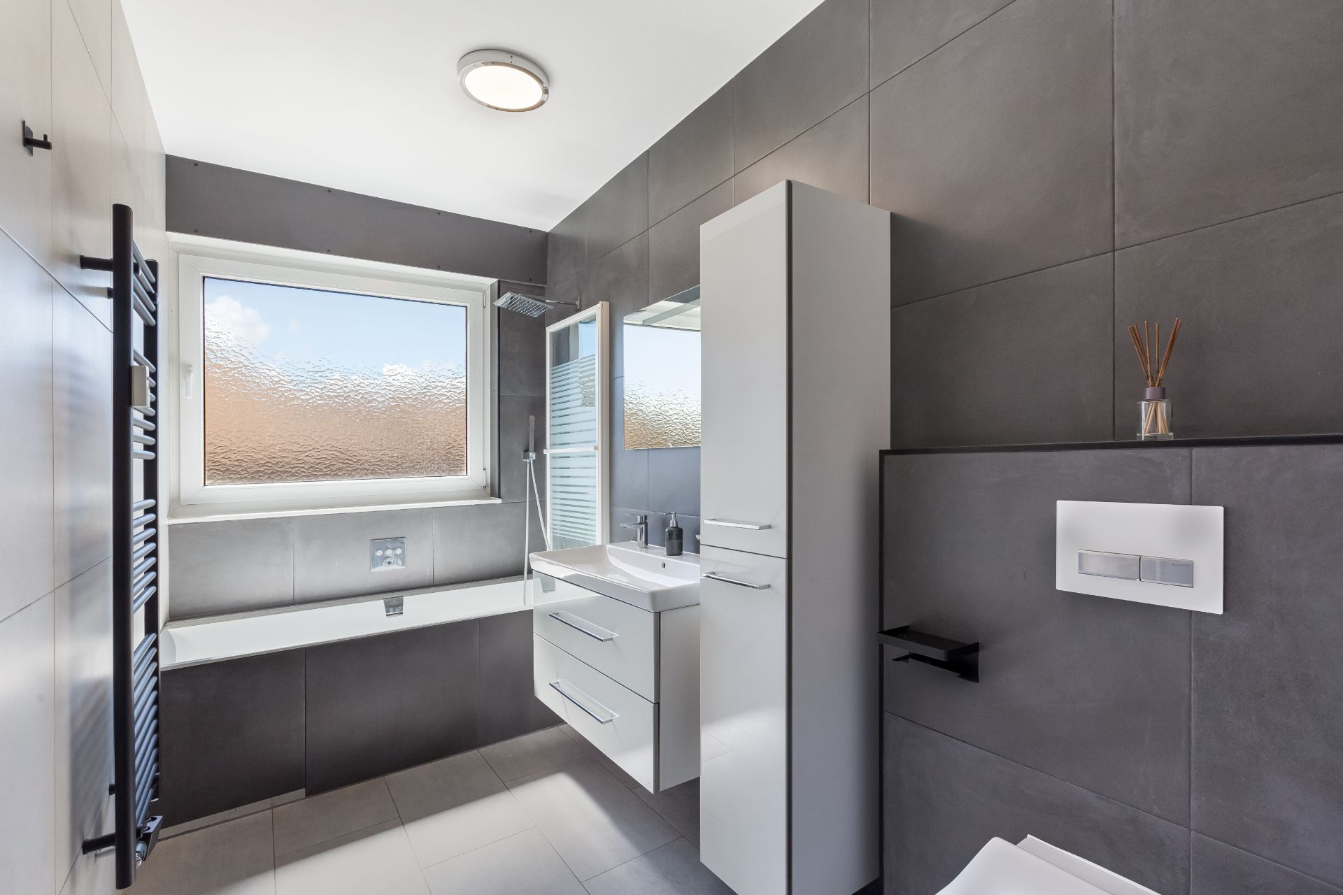 Modernes Badezimmer mit Badewanne, großem Fenster und grauen Fliesen.