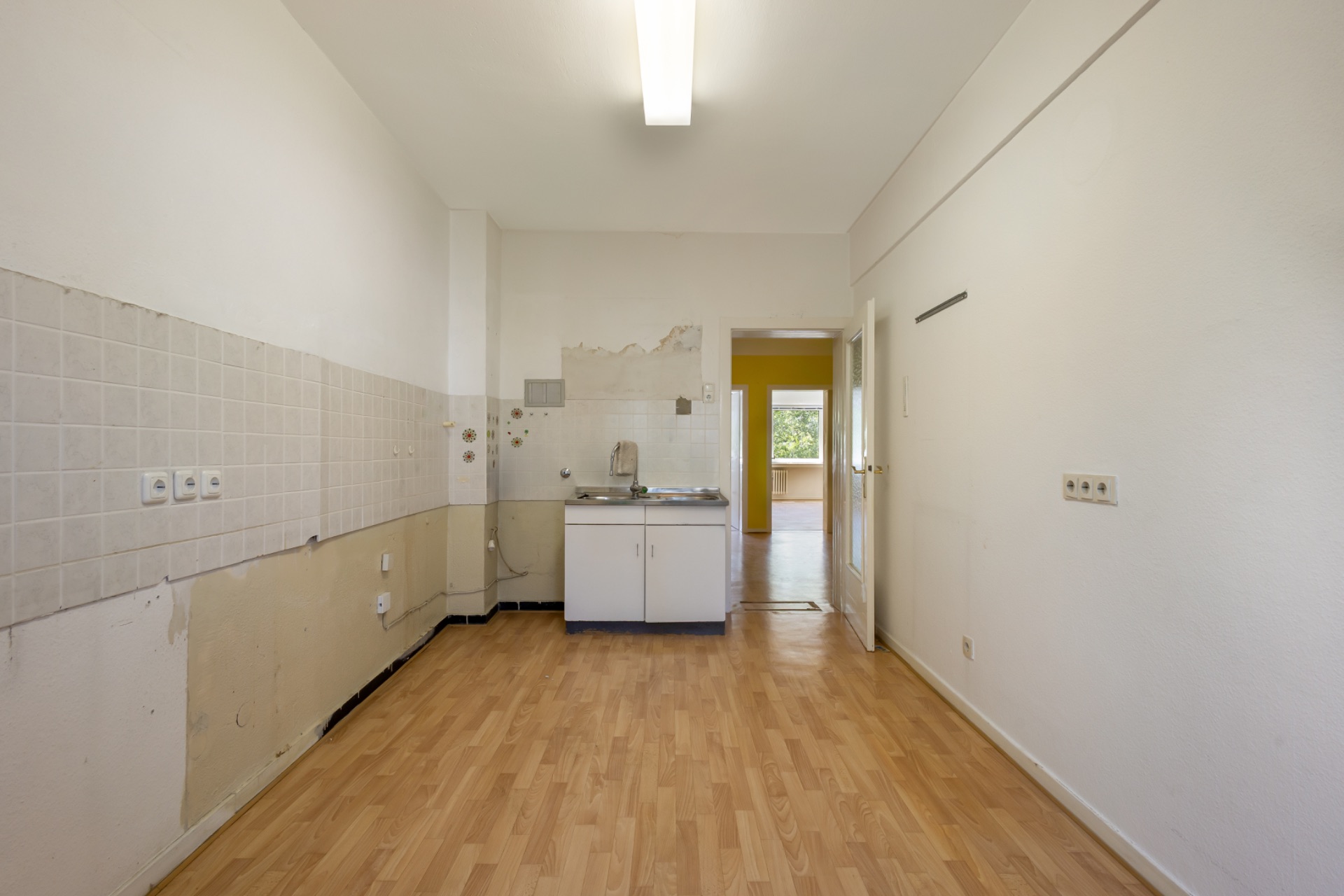 Leerer Küchenraum mit Holzboden, Spüle und Zugang zu einem anderen Zimmer.
