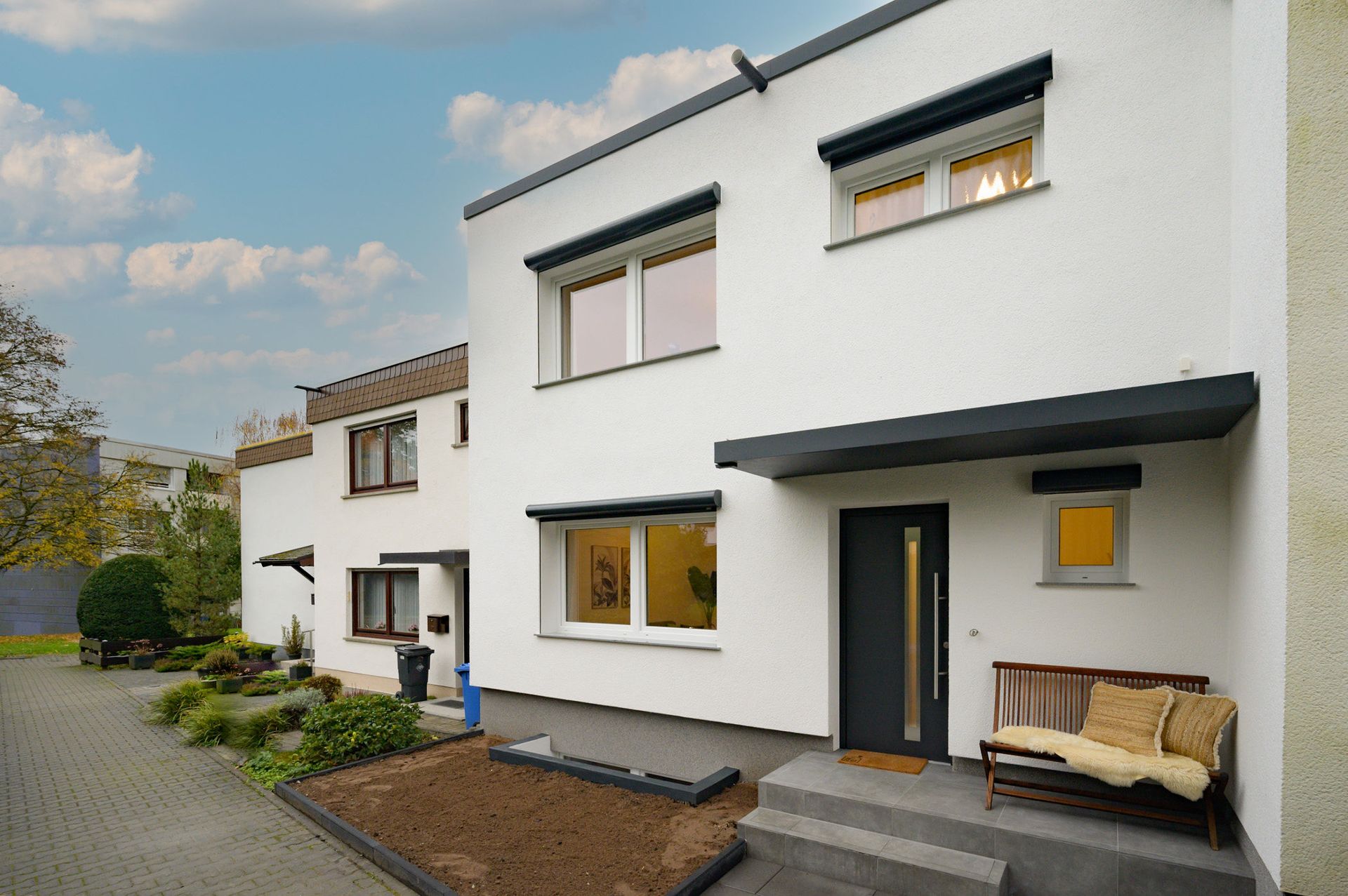 Moderne weiße Hausfassade mit Eingangsbereich, Bank und kleinen Gartenflächen.