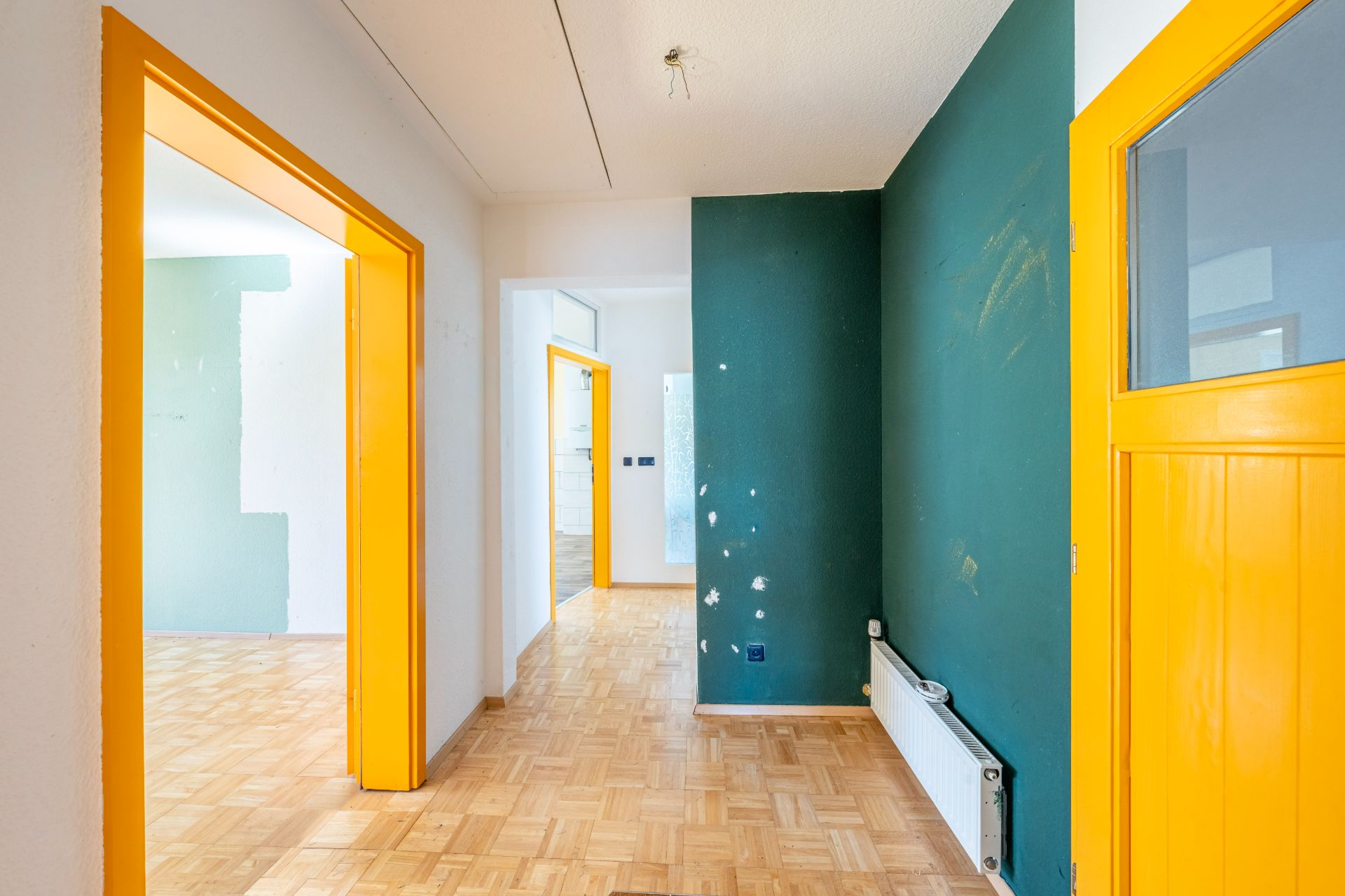 Flur mit grün gestrichener Wand, gelben Türrahmen und Parkettboden in einer Wohnung.