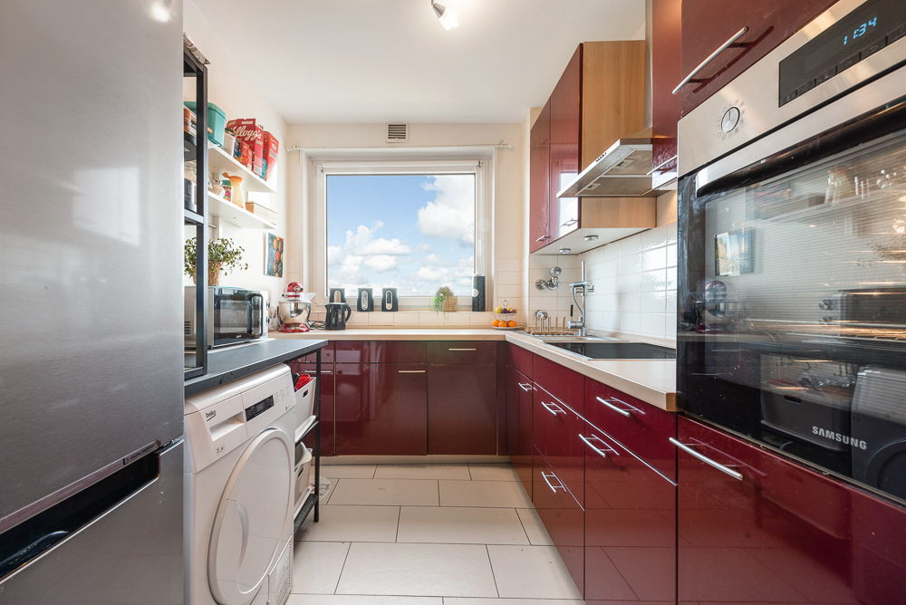 Moderne Küche mit roten Schränken, weißen Geräten und großem Fenster mit Blick nach draußen.
