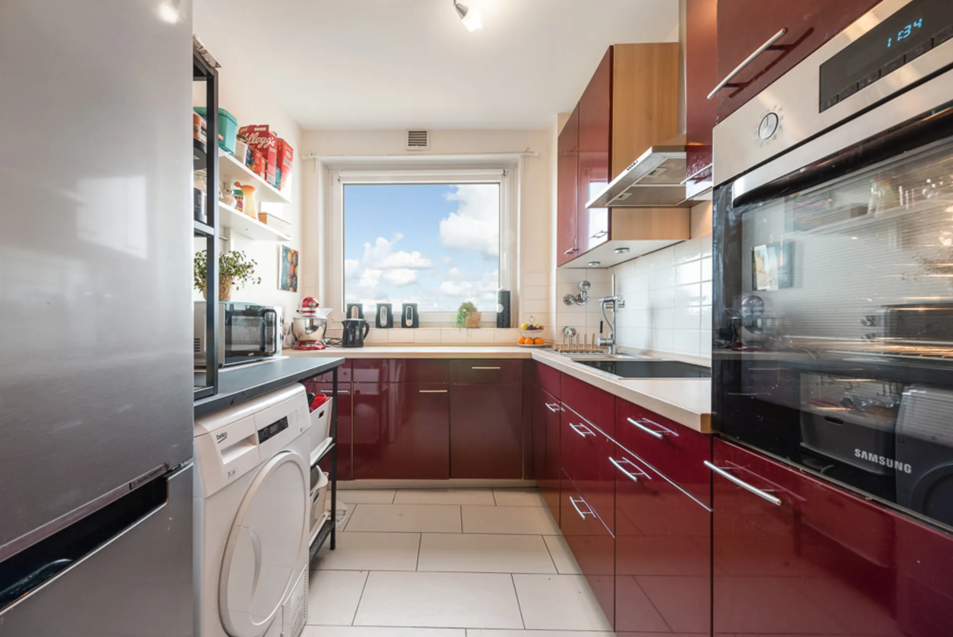 Moderne Küche mit roten Schränken, weißen Geräten und großem Fenster mit Blick nach draußen.