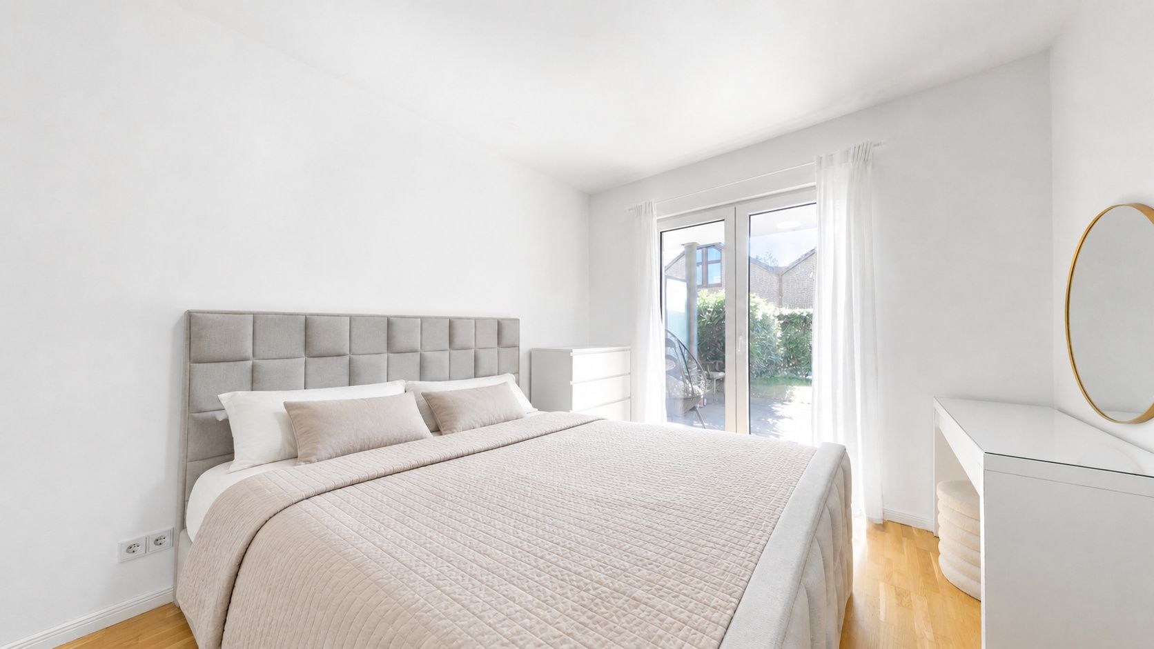 Helles Schlafzimmer mit großem Bett, Kommode, Spiegel und Fenster mit Blick nach draußen.