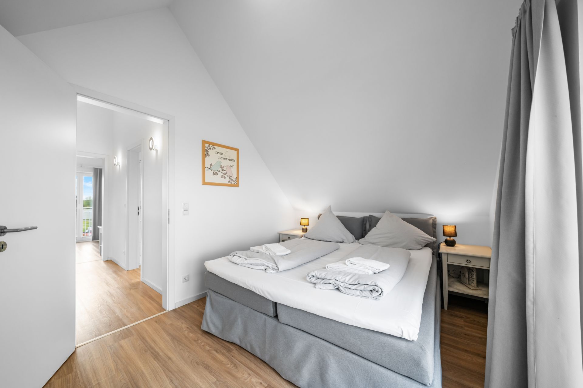 Helles Schlafzimmer mit Doppelbett, zwei Nachttischen und offenem Durchgang zu einem Flur.