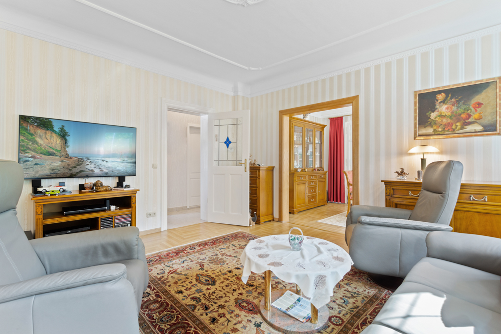 Helles Wohnzimmer mit Teppich, zwei grauen Sesseln, Fernseher und Holzmöbeln.