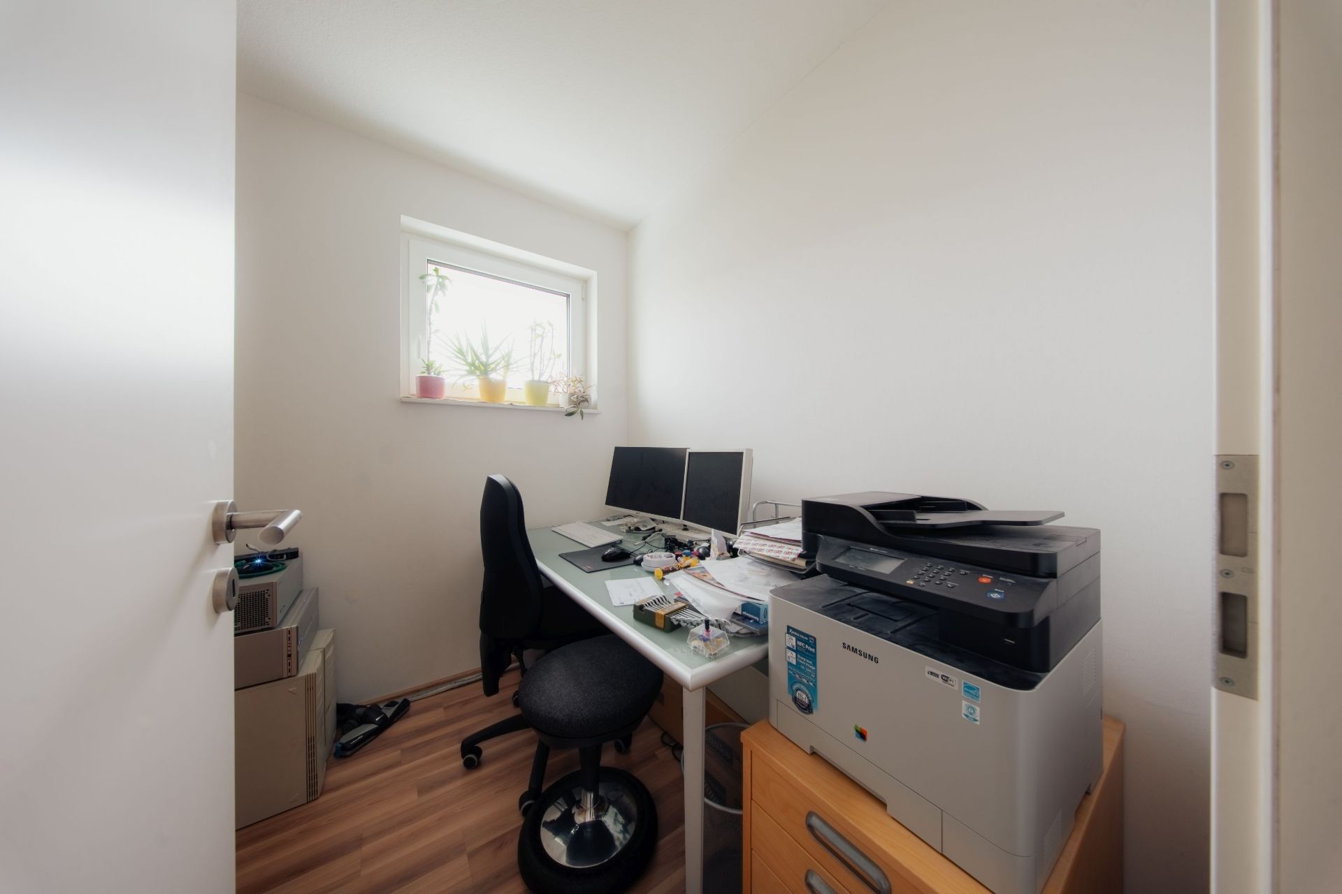 Kleines Arbeitszimmer mit Schreibtisch, zwei Monitoren, Drucker und Pflanzen auf der Fensterbank.