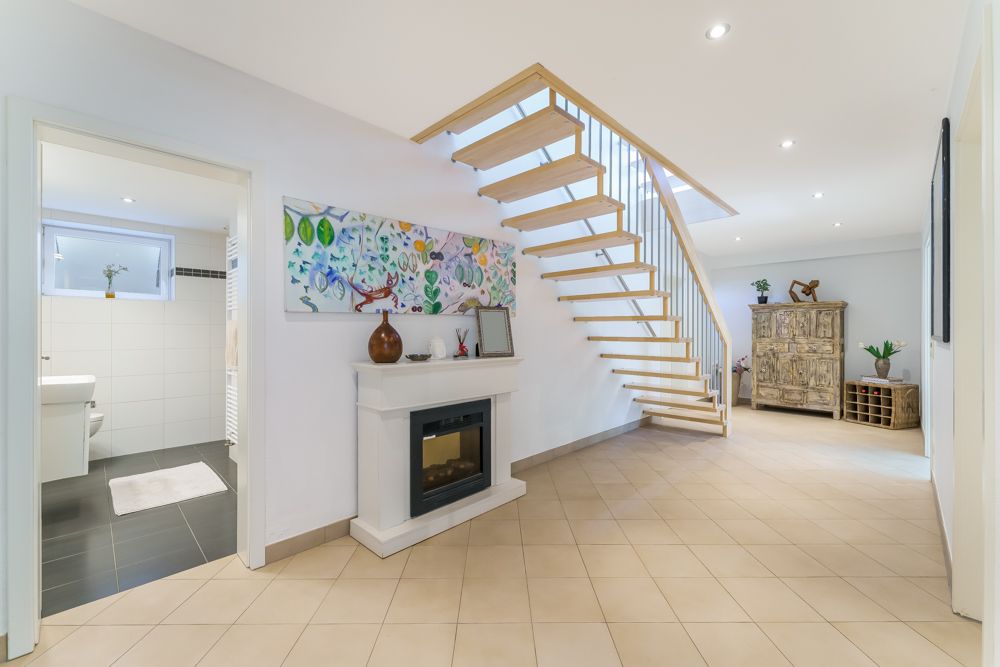 Heller Flur mit offener Holztreppe, Kamin und Zugang zu einem Badezimmer, im Hintergrund ein Sideboard.