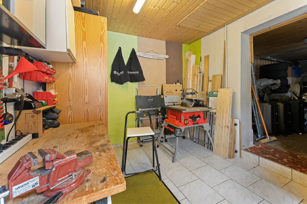 Werkstatt mit Holztisch, Schraubstock, Werkzeugen und Brettern auf weißem Fliesenboden.