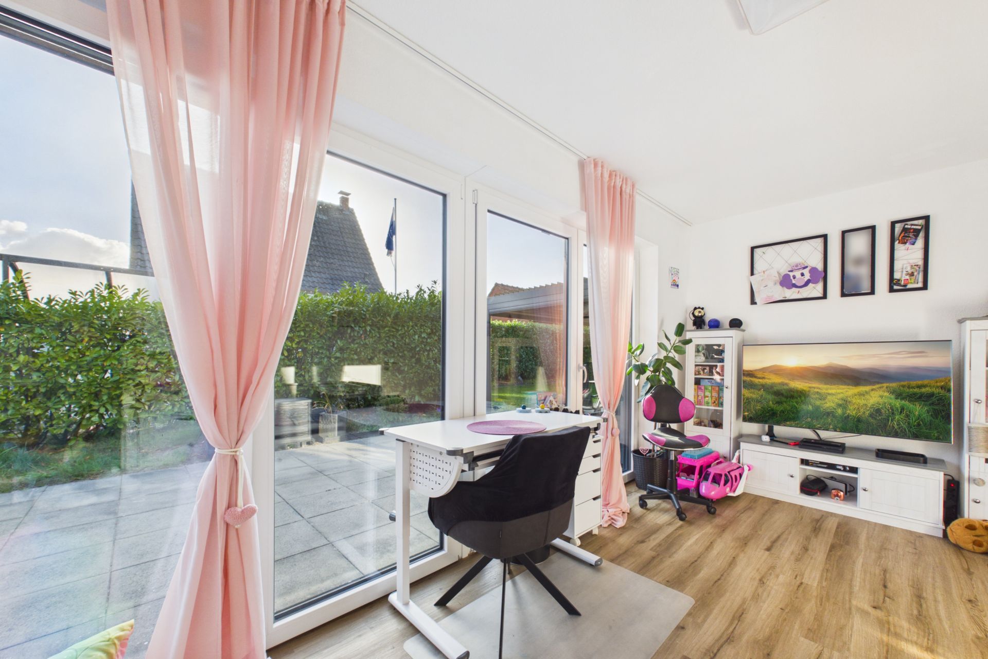 Helles Wohnzimmer mit Schreibtisch, Fernseher und großen Fenstern mit rosa Vorhängen.