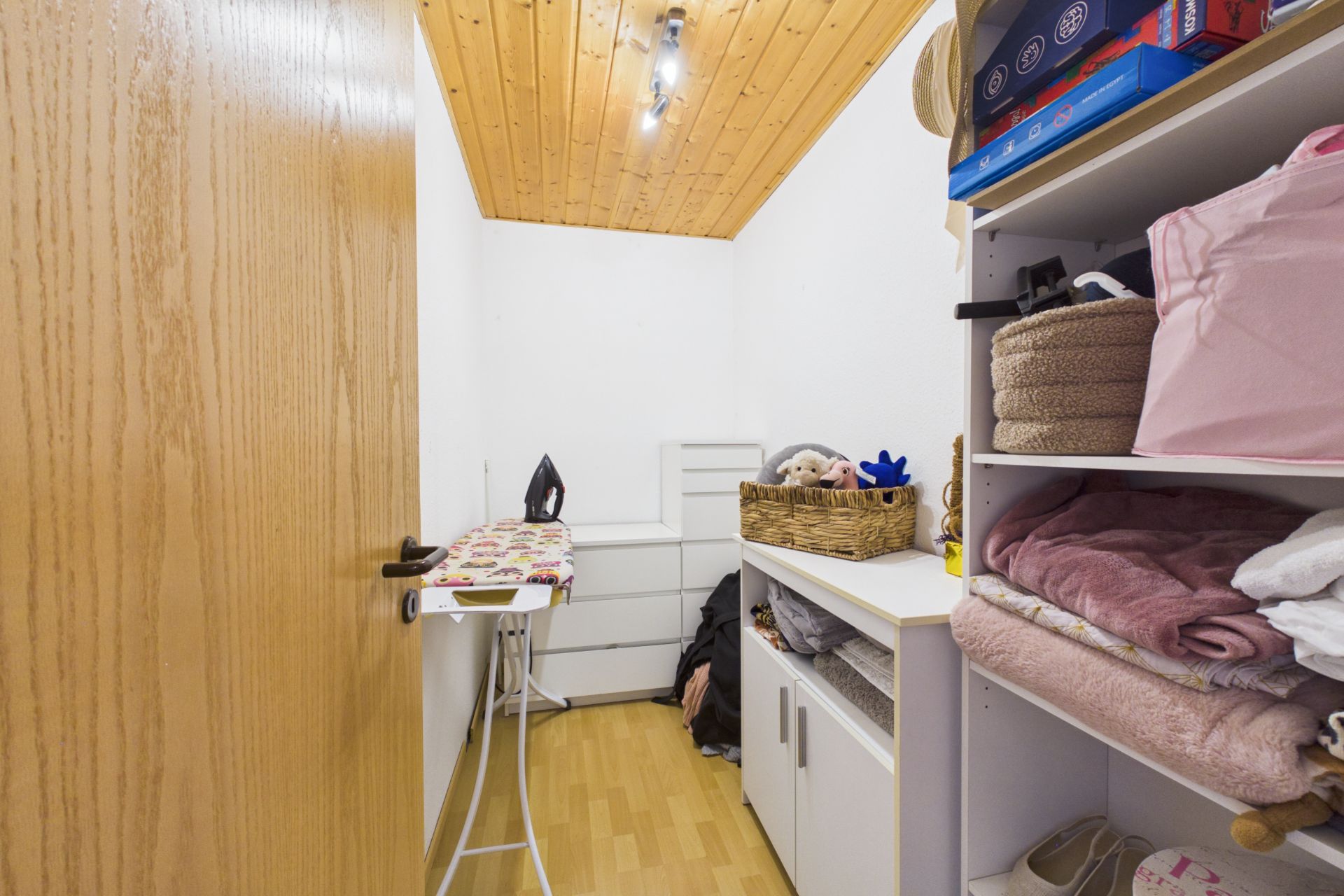 Kleiner Hauswirtschaftsraum mit Regal, Kommode, Bügelbrett und Holzdecke.