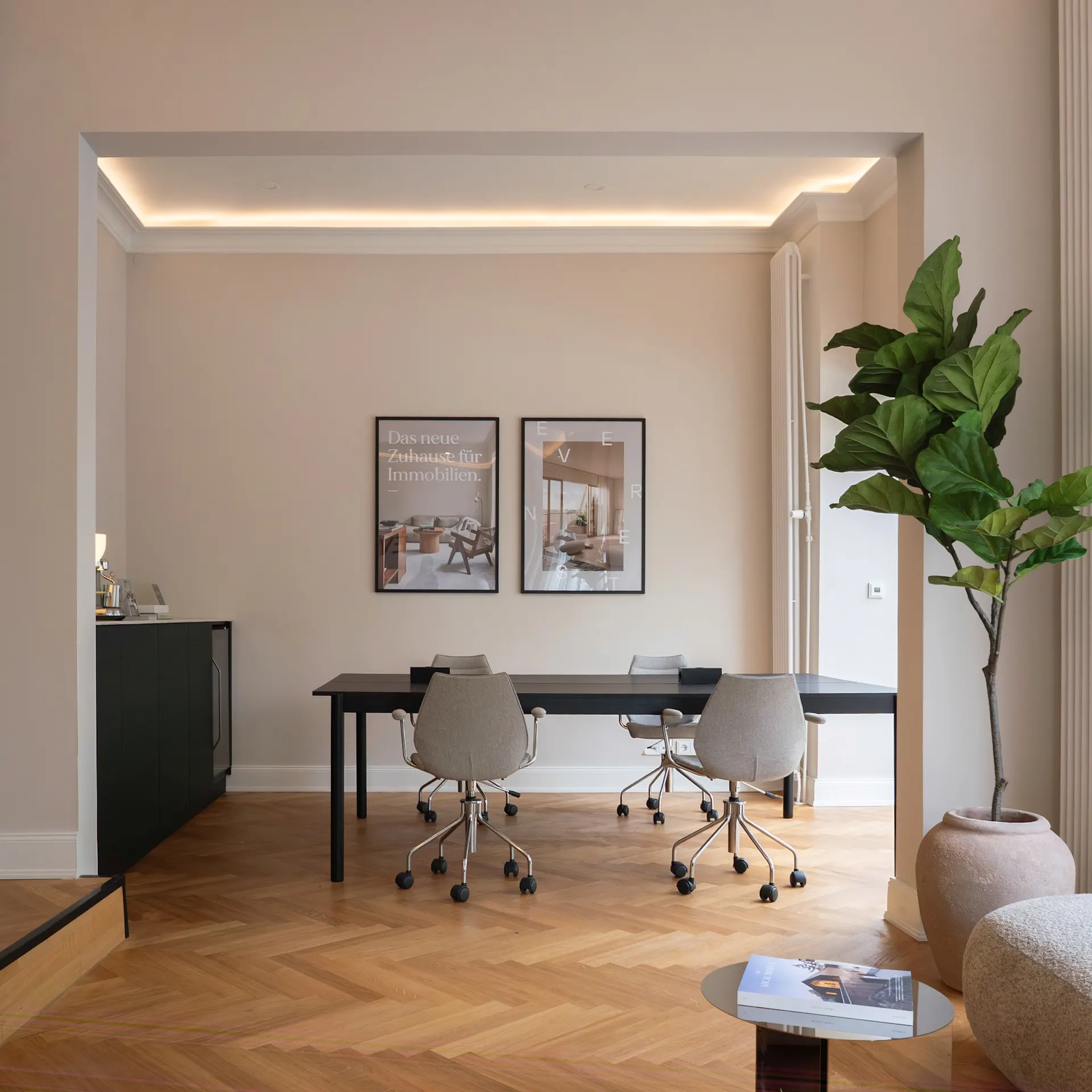 Heller Raum mit Holzboden, Schreibtisch, drei Stühlen, großer Pflanze und zwei Bildern an der Wand.