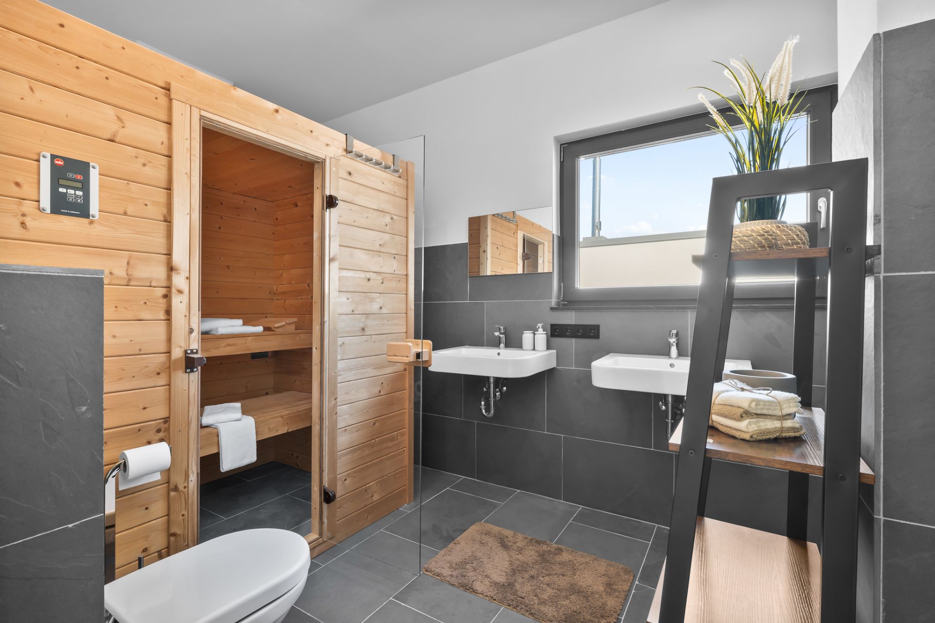 Modernes Badezimmer mit Sauna, zwei Waschbecken, Regal, WC und großem Fenster.