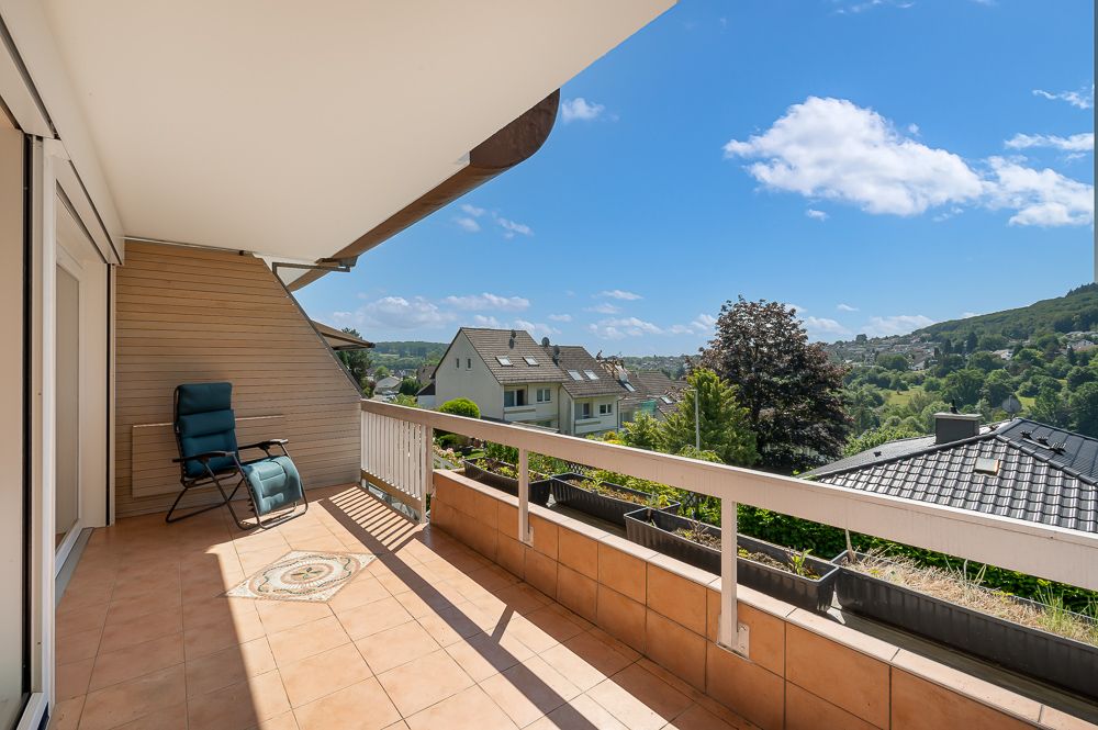 Geräumiger Balkon mit Stuhl, Fliesenboden und Ausblick auf Wohnhäuser und grüne Hügel.