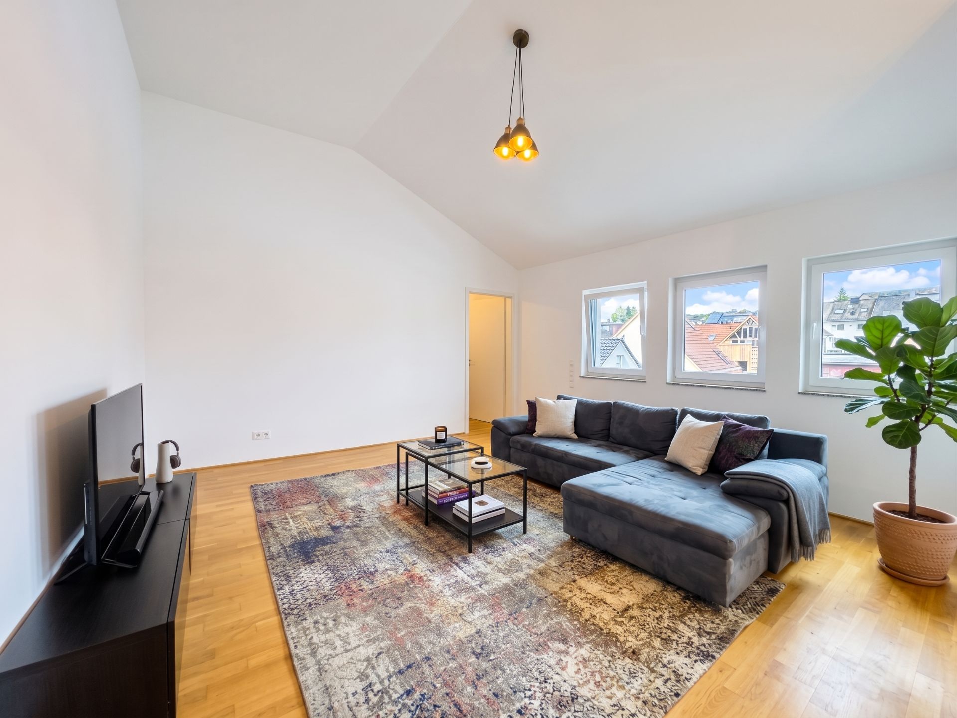 Helles Wohnzimmer mit Sofa, Teppich, Fernseher, Couchtisch und Pflanze vor drei Fenstern.