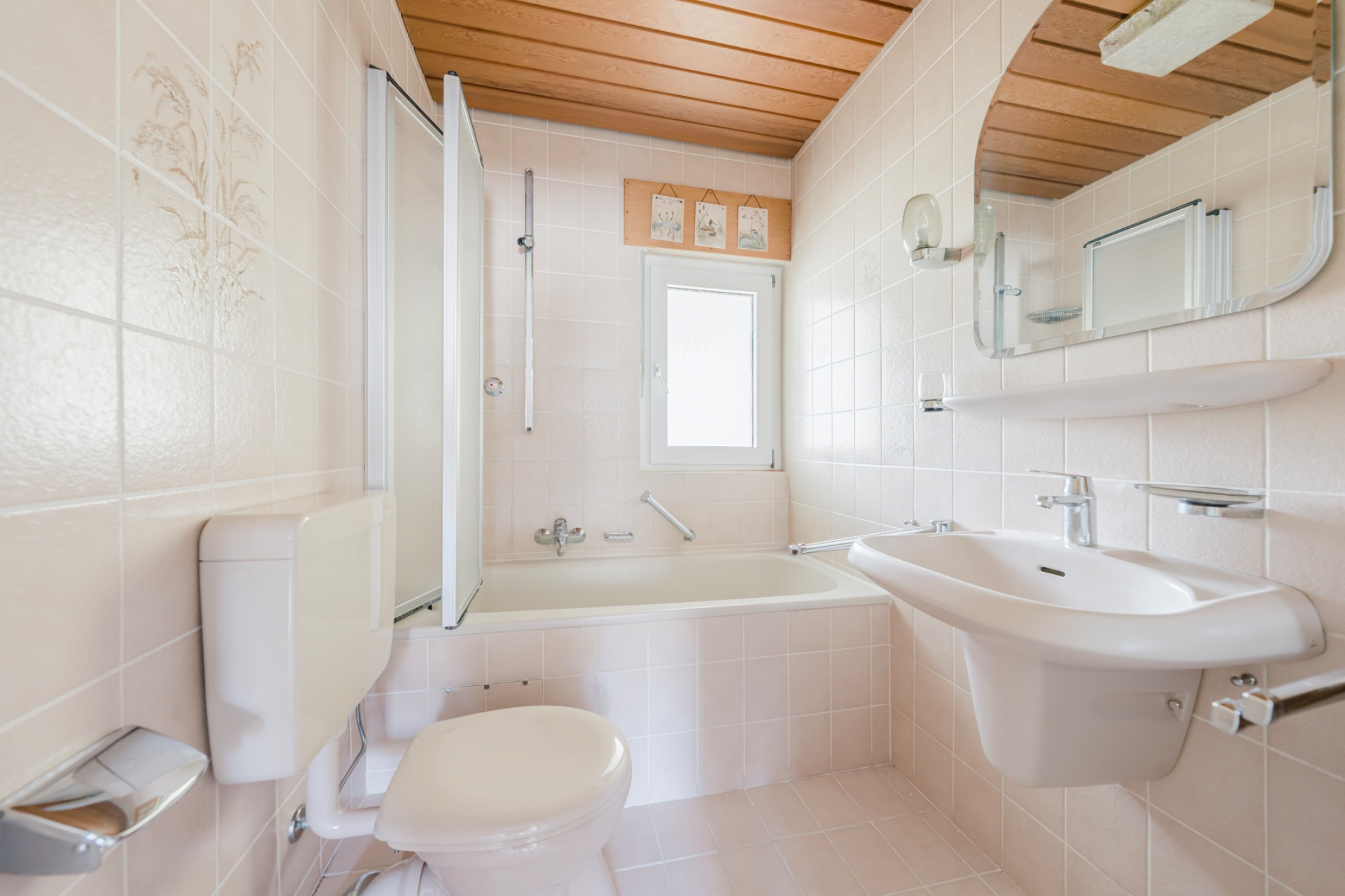 Helles Badezimmer mit Badewanne, Waschbecken, Toilette und Fenster, Wandspiegel und Holzdecke.