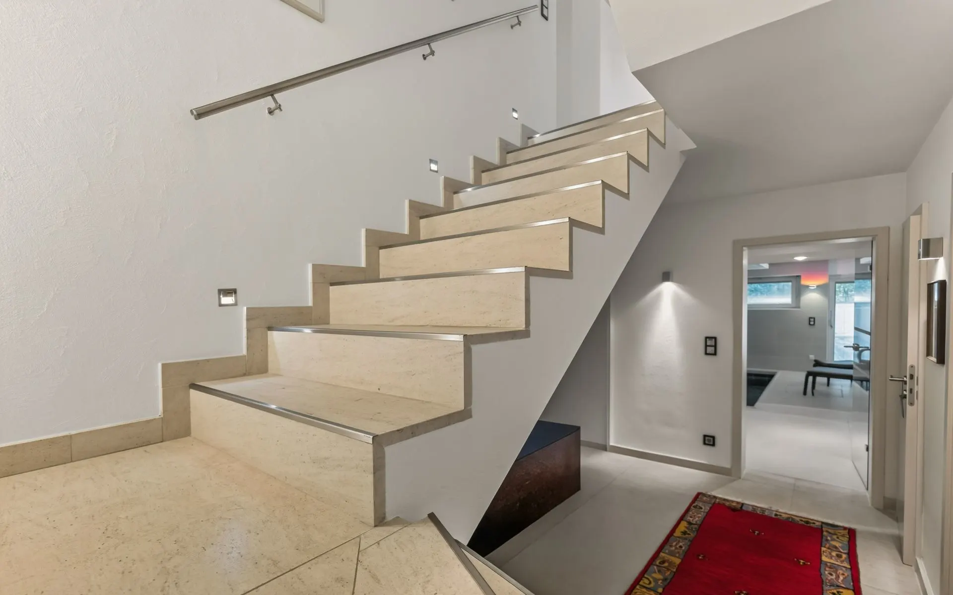 Heller Flur mit Treppe aus hellem Stein und rotem Teppich, Blick in ein Wohnzimmer.