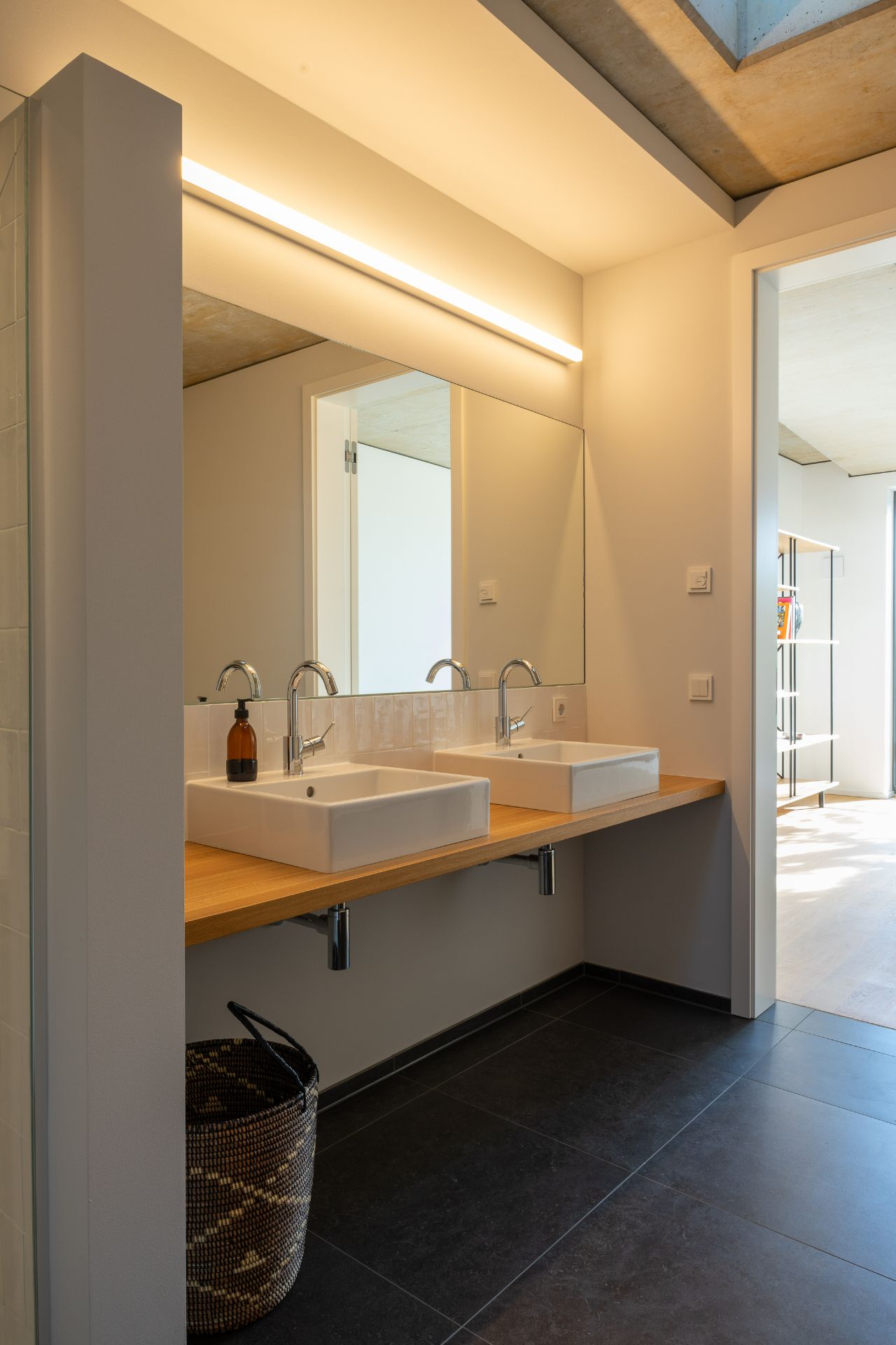Modernes Badezimmer mit zwei Waschbecken, großem Spiegel und Wäschekorb auf dunklem Fliesenboden.