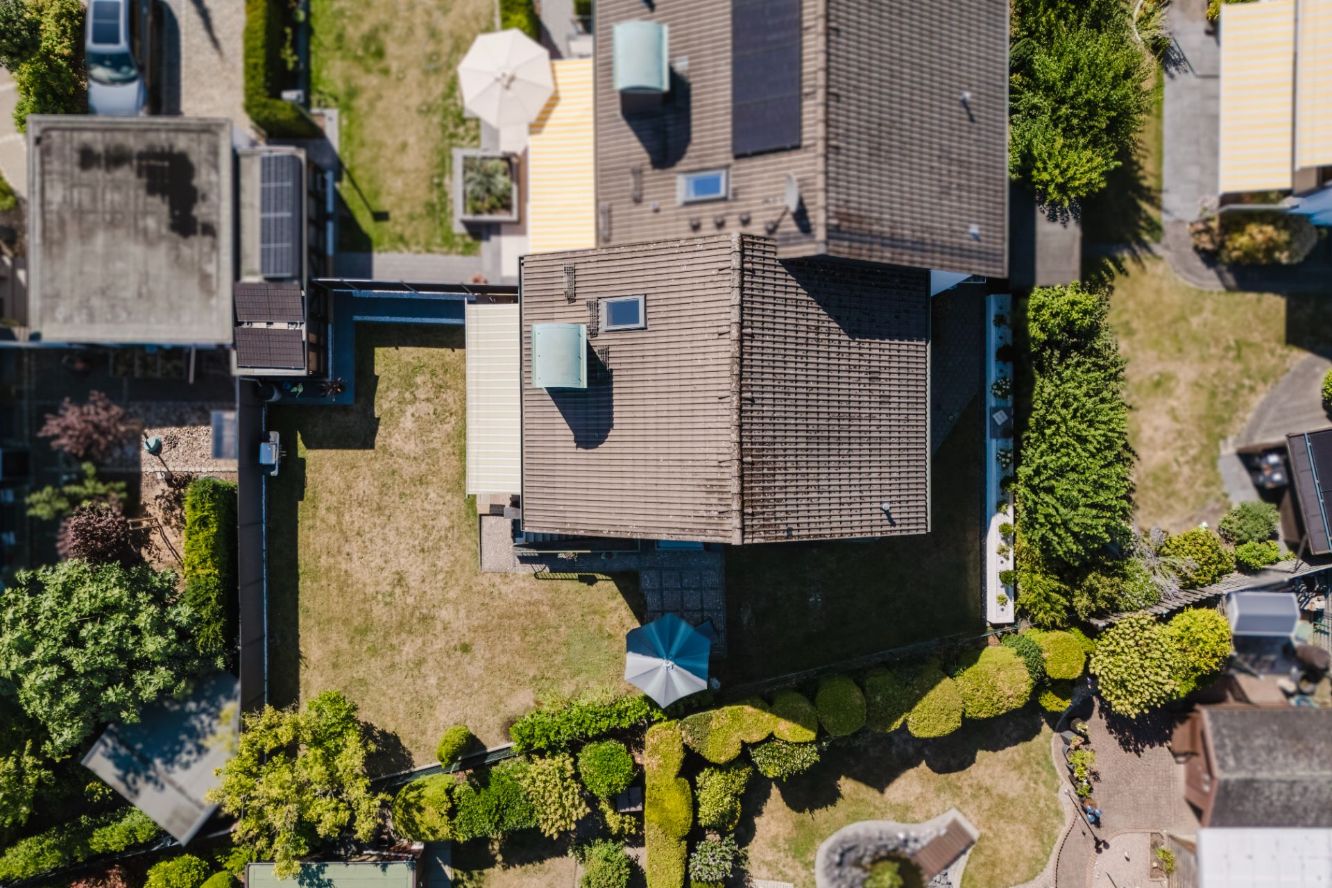 Luftaufnahme eines Hauses mit Garten, Terrasse und Sonnenschirm, umgeben von Hecken und Nachbargrundstücken.