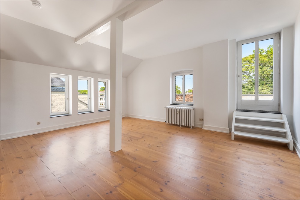 Helles, leeres Zimmer mit Holzboden, mehreren Fenstern und einer Tür mit Stufen nach draußen.